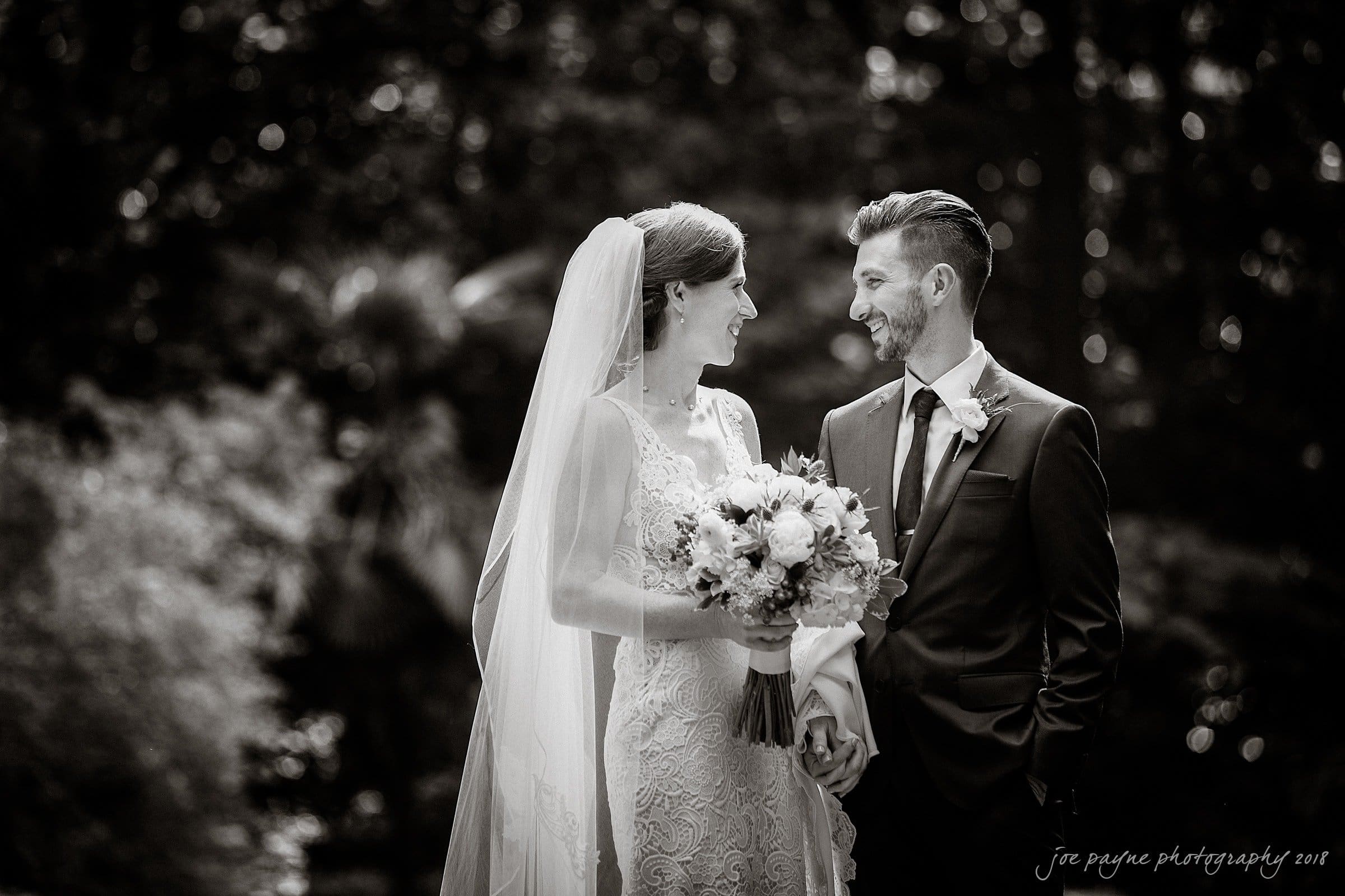 A black and white photo of a smiling couple on their wedding day, holding hands. The bride is in a lacy gown with a veil, holding a bouquet. The groom is in a dark suit with a boutonnière. They are looking at each other lovingly.