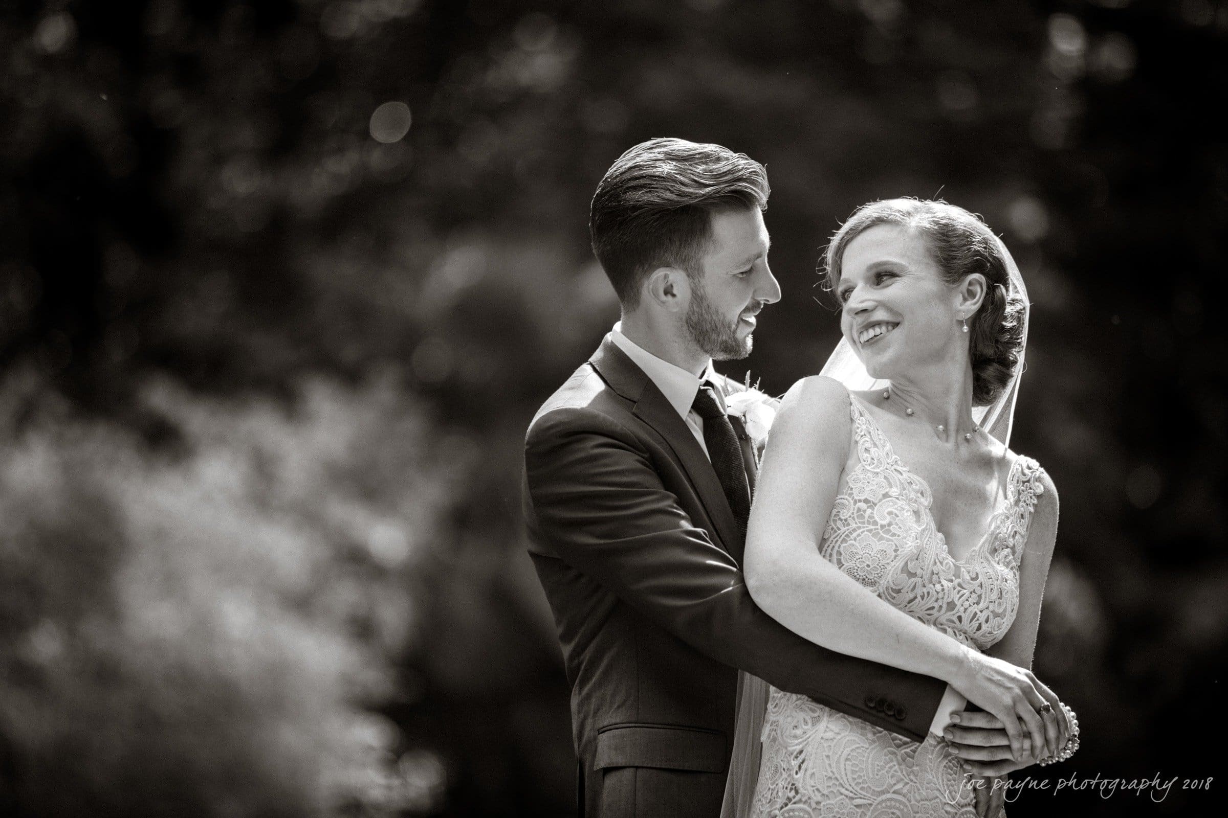 A black and white photo of a couple on their wedding day. They are embracing and smiling at each other. The groom is in a suit and the bride is wearing a lace wedding dress with a veil. The background is softly blurred, creating a romantic atmosphere.