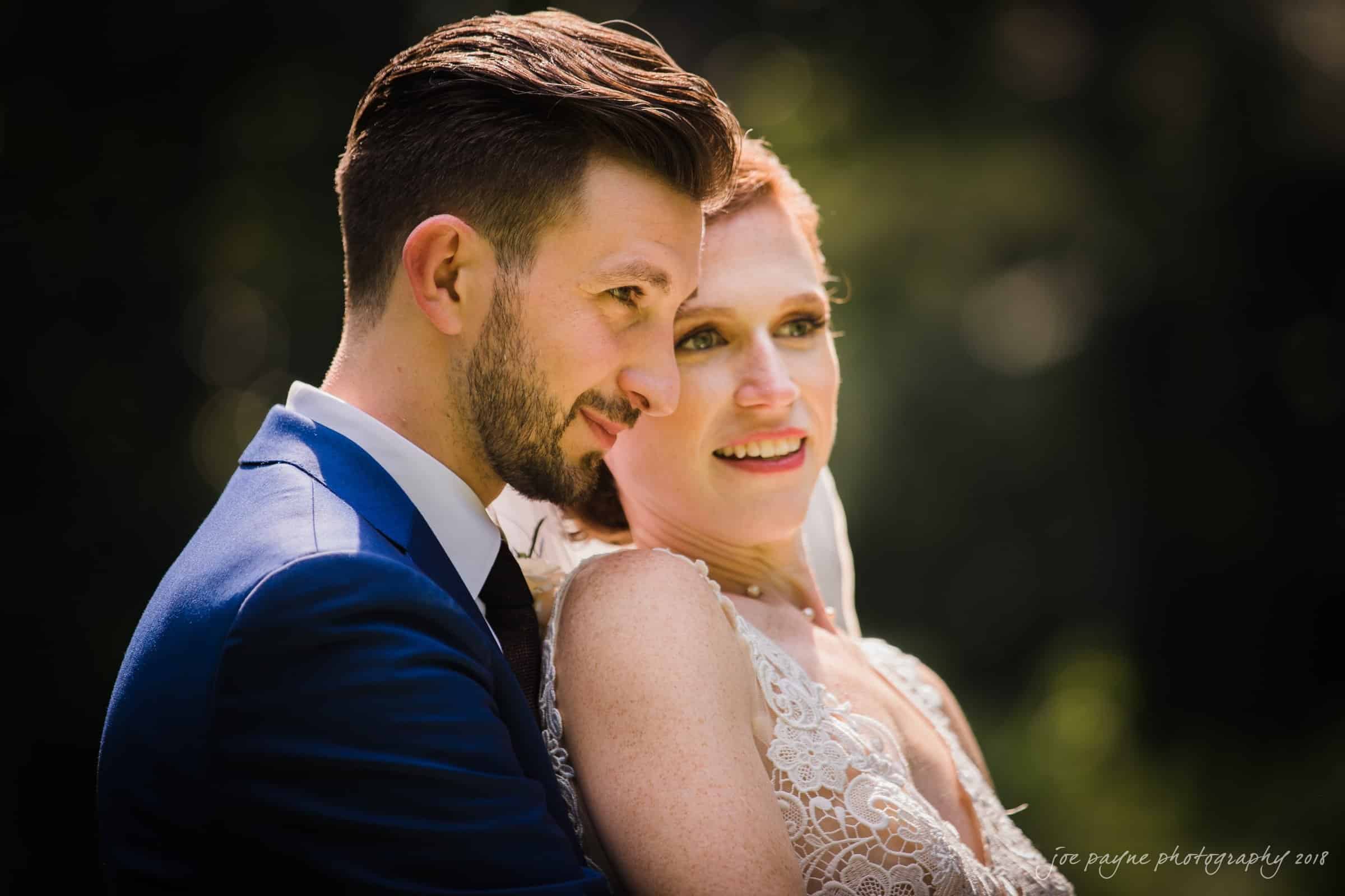 A bride and groom share an intimate, joyful moment outdoors. The groom, in a blue suit, embraces the bride, who is wearing a lacy white dress. Sunlight highlights their expressions against a blurred natural background.