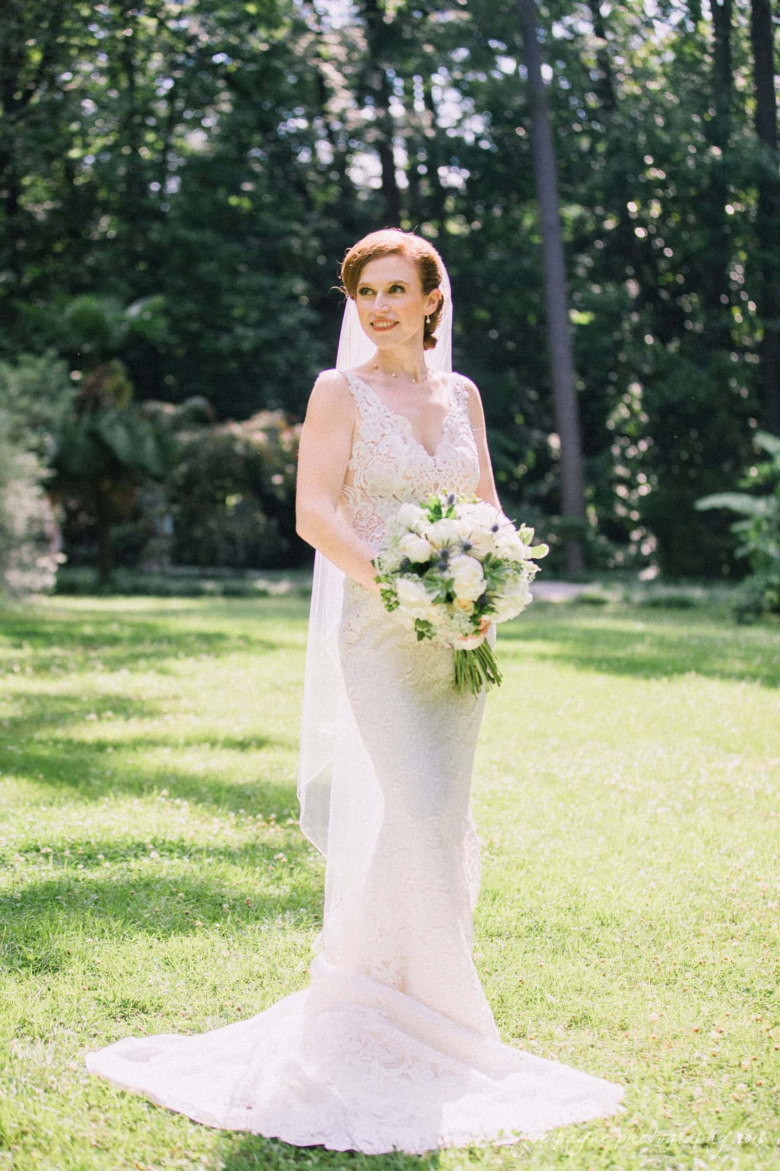 A bride stands on a sunlit, grassy field, wearing a white lace gown and veil. She holds a bouquet of white and green flowers, smiling softly. Tall trees provide a lush, verdant backdrop.