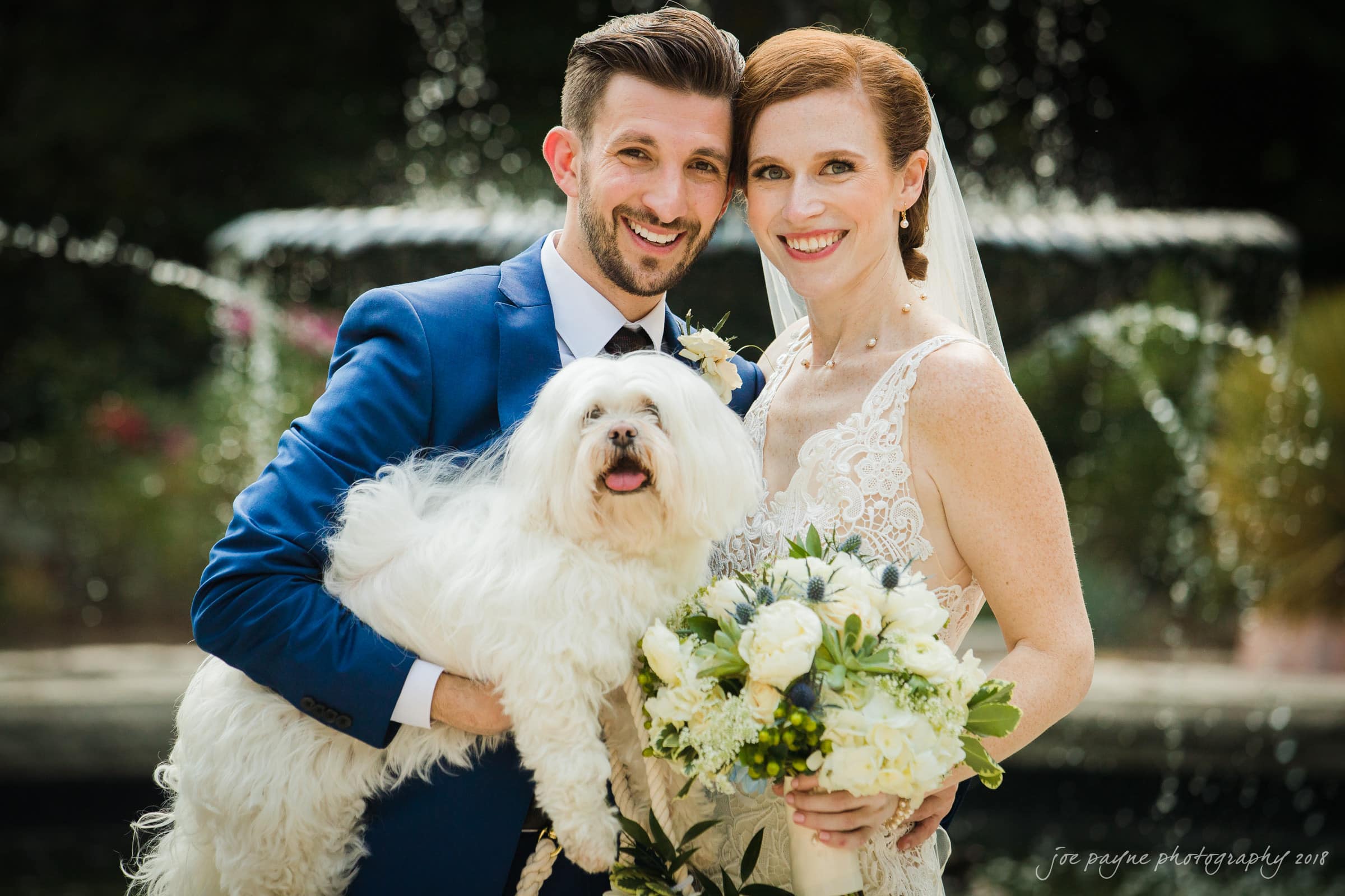 A bride and groom smile while posing with a fluffy white dog. The groom is in a blue suit, and the bride is in a white dress holding a bouquet. A fountain is blurred in the background.