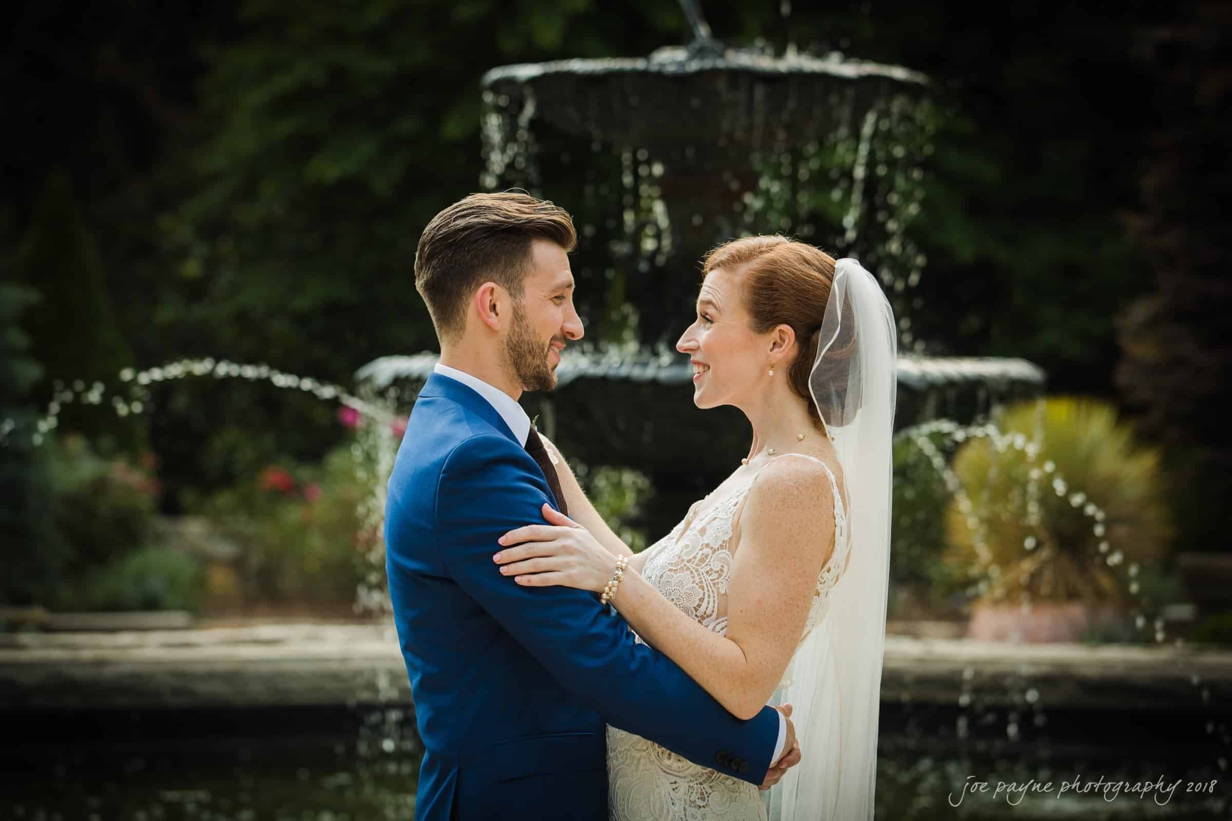 A bride and groom face each other, smiling in front of a large, elegant fountain. The groom wears a blue suit, and the bride is in a lace wedding dress with a veil. Lush greenery surrounds them.