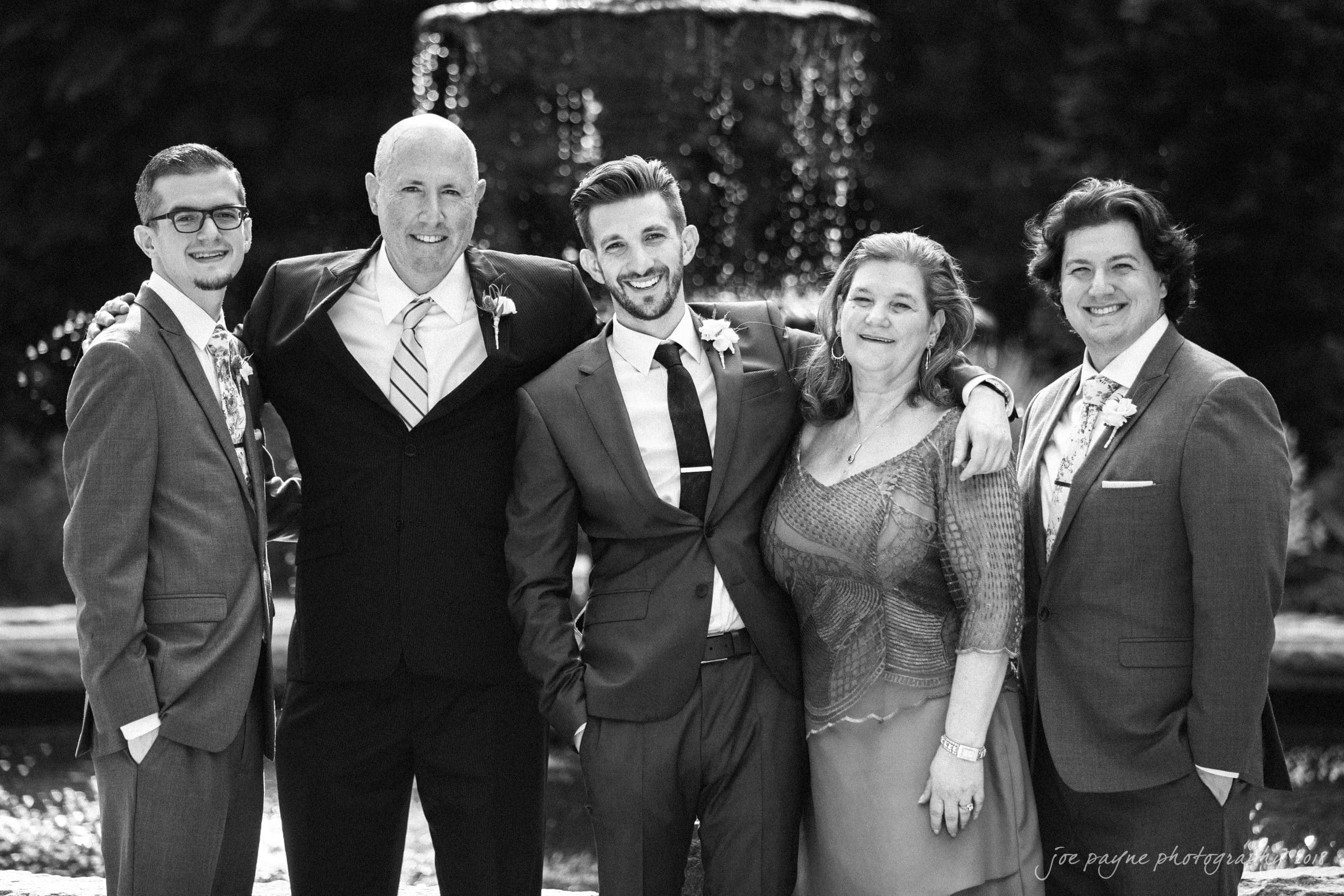 A group of five people posing in front of a fountain. All are dressed in formal attire, with the men wearing suits and the woman wearing a formal dress. They are smiling and standing closely together, conveying a sense of camaraderie.
