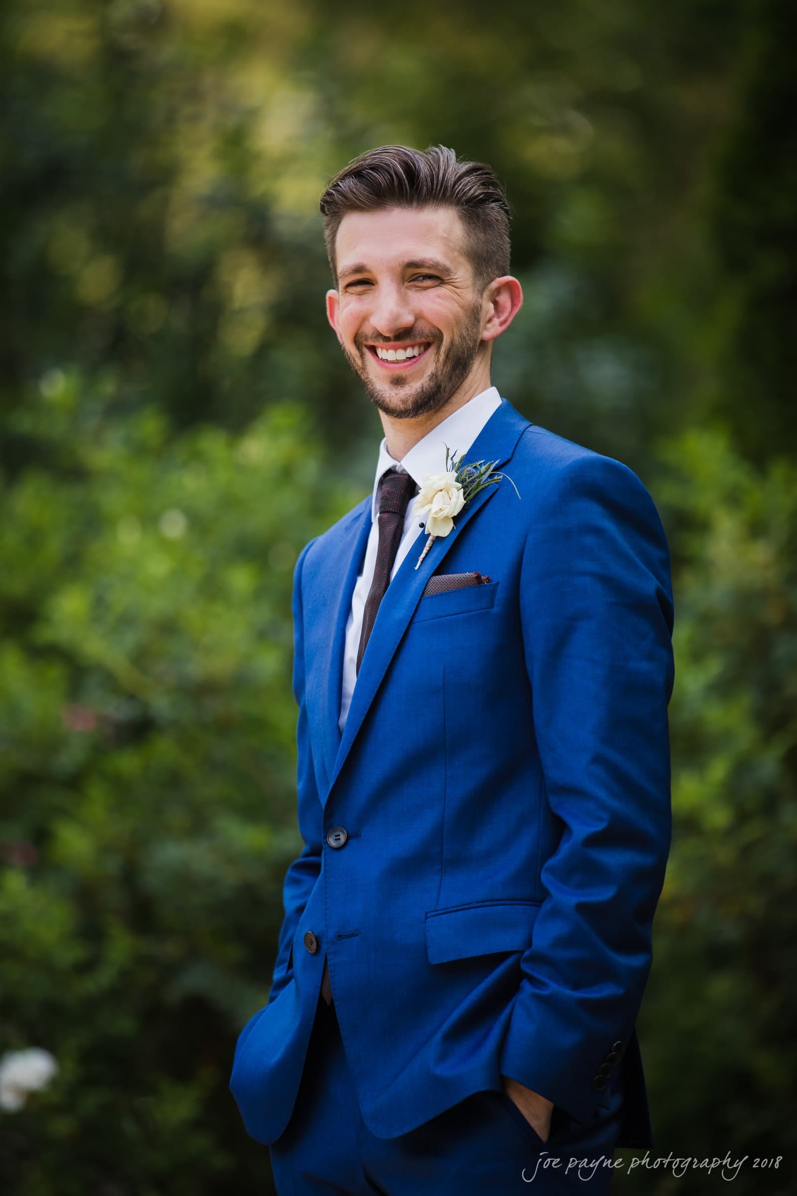 A man in a bright blue suit with a white boutonniere smiles while standing in a garden. His hands are in his pockets, and he wears a white shirt with a dark tie. The background is blurred greenery.