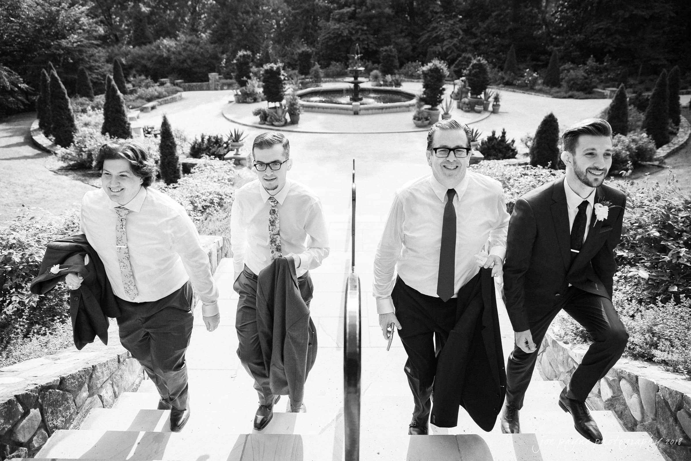 Four men in formal attire, three in shirts and ties holding jackets, and one in a suit, walk up steps in a garden. A fountain and trees are visible in the background. The image is in black and white.