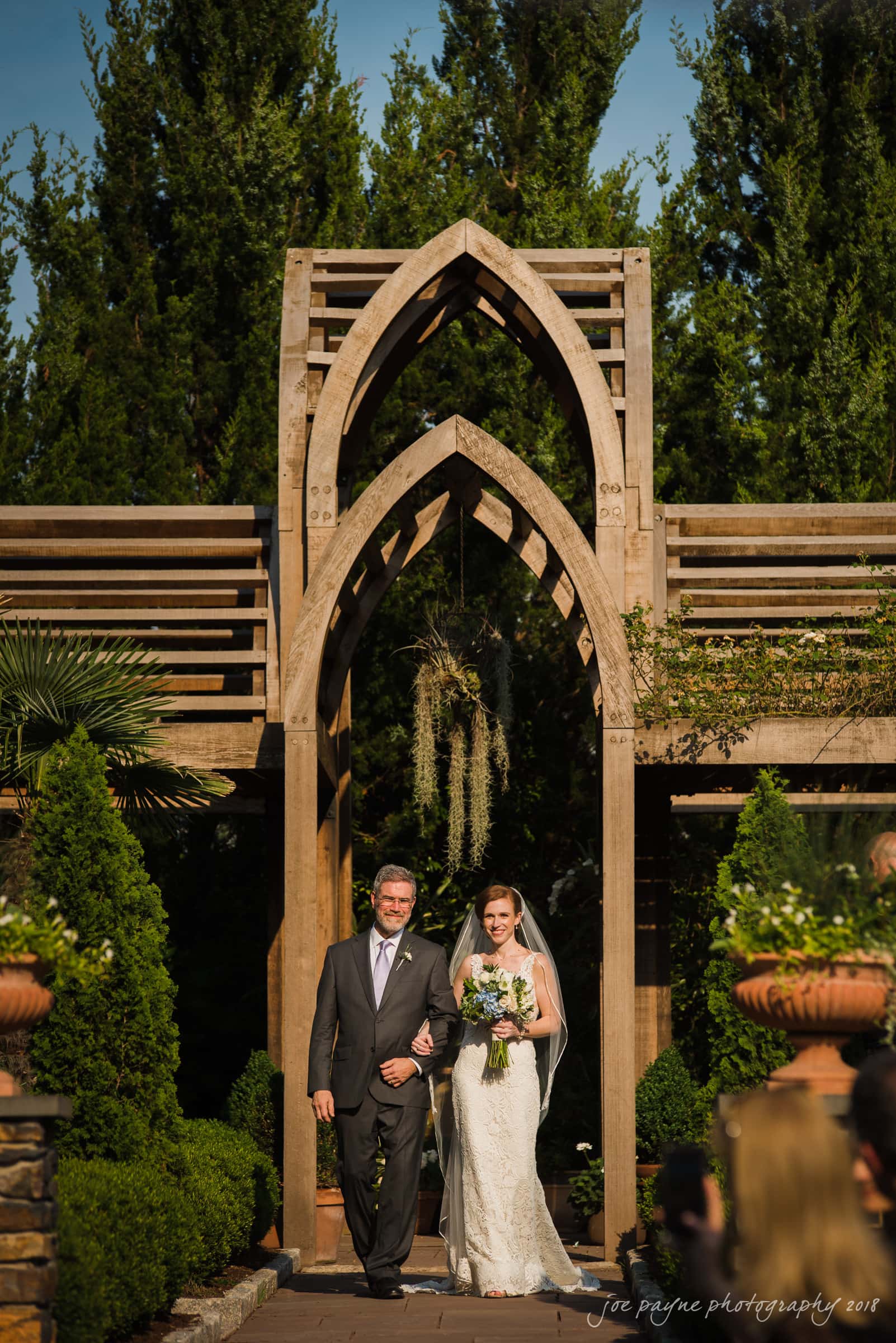 A bride in a white dress and veil walks arm-in-arm with a man in a suit under a wooden archway in a garden setting. Both are smiling, surrounded by greenery and potted plants.