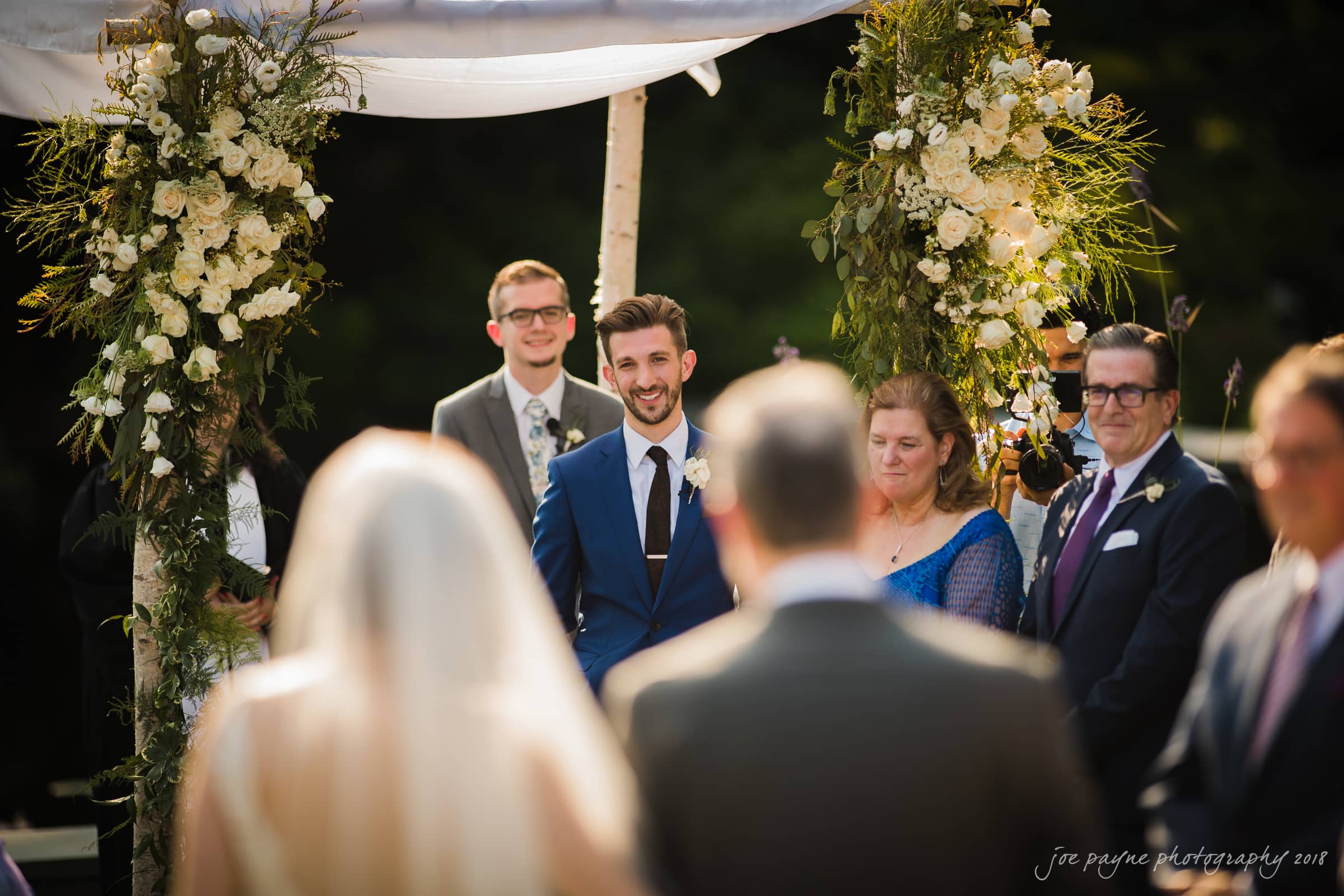 A wedding ceremony scene with a bride and groom standing under a decorated floral arch. The groom smiles, wearing a blue suit, while guests in formal attire watch. The setting is outdoors, with greenery in the background.