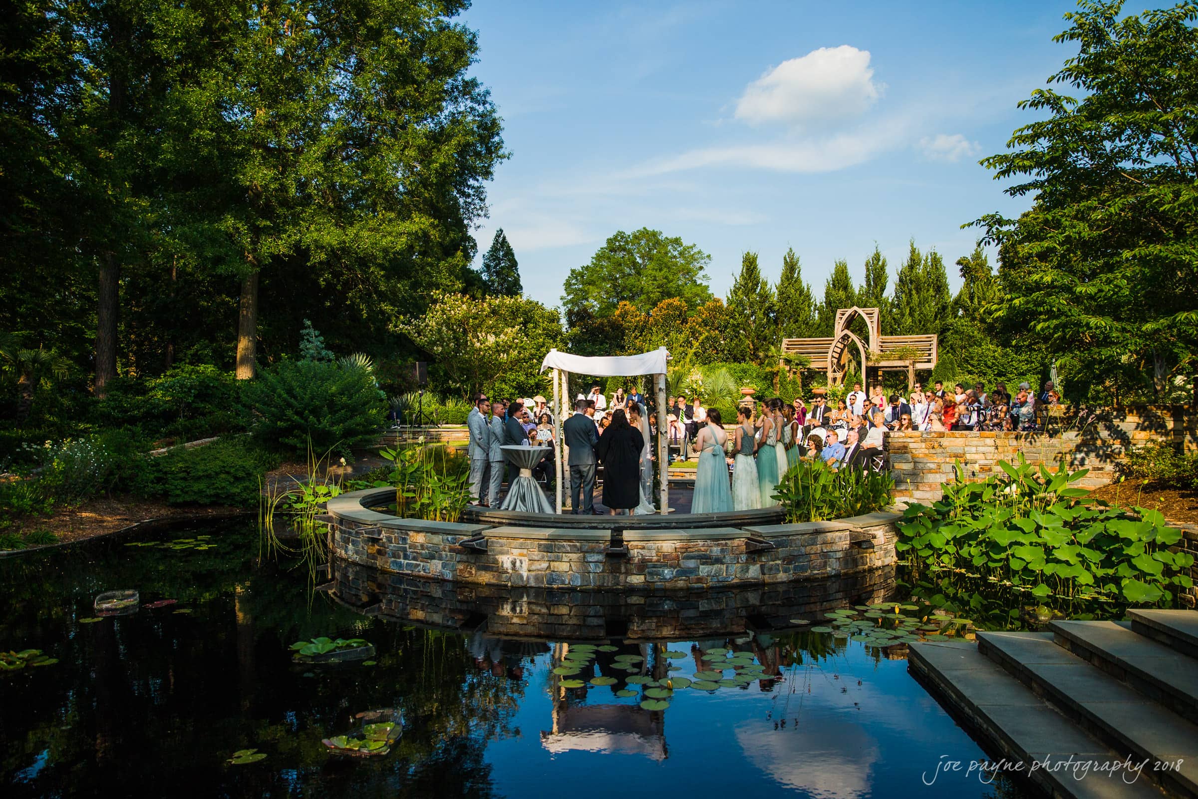 A wedding ceremony takes place on a stone platform surrounded by a pond and lush greenery. Guests are seated around the structure, and the sky is clear and blue. A white canopy is set up above the couple at the altar.