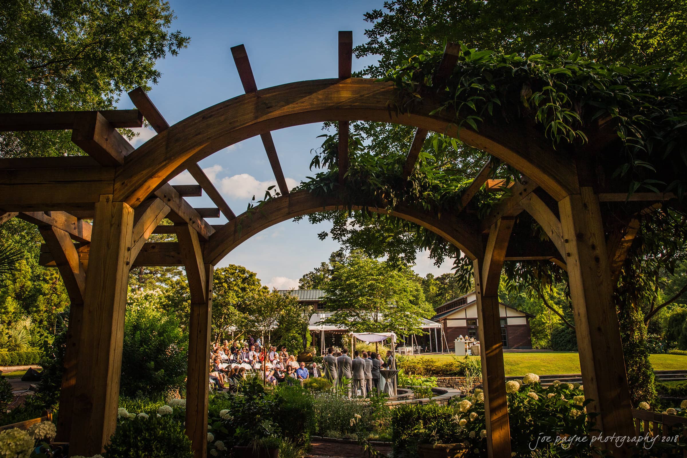 A lush outdoor garden scene under a wooden archway. Guests are seated for a wedding ceremony, facing an altar adorned with white drapes. Sunlight filters through trees, creating a vibrant summer ambiance.
