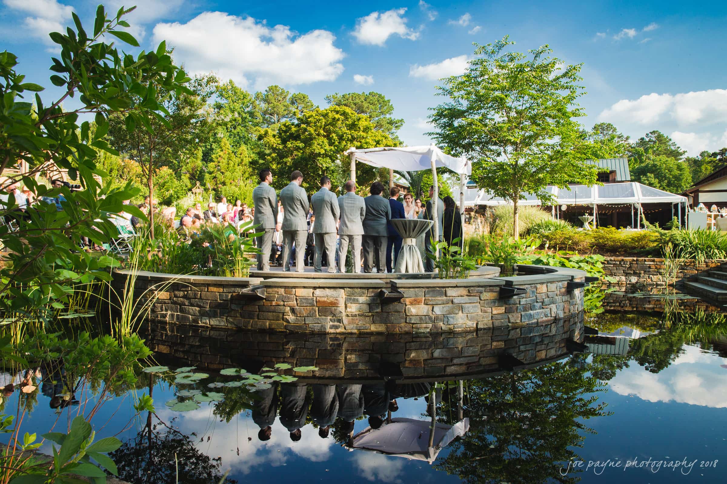 A wedding ceremony takes place on a circular stone platform surrounded by a pond with lily pads. Guests watch under a clear blue sky, with lush greenery in the background. Bridesmaids and groomsmen stand beside the couple under a white canopy.