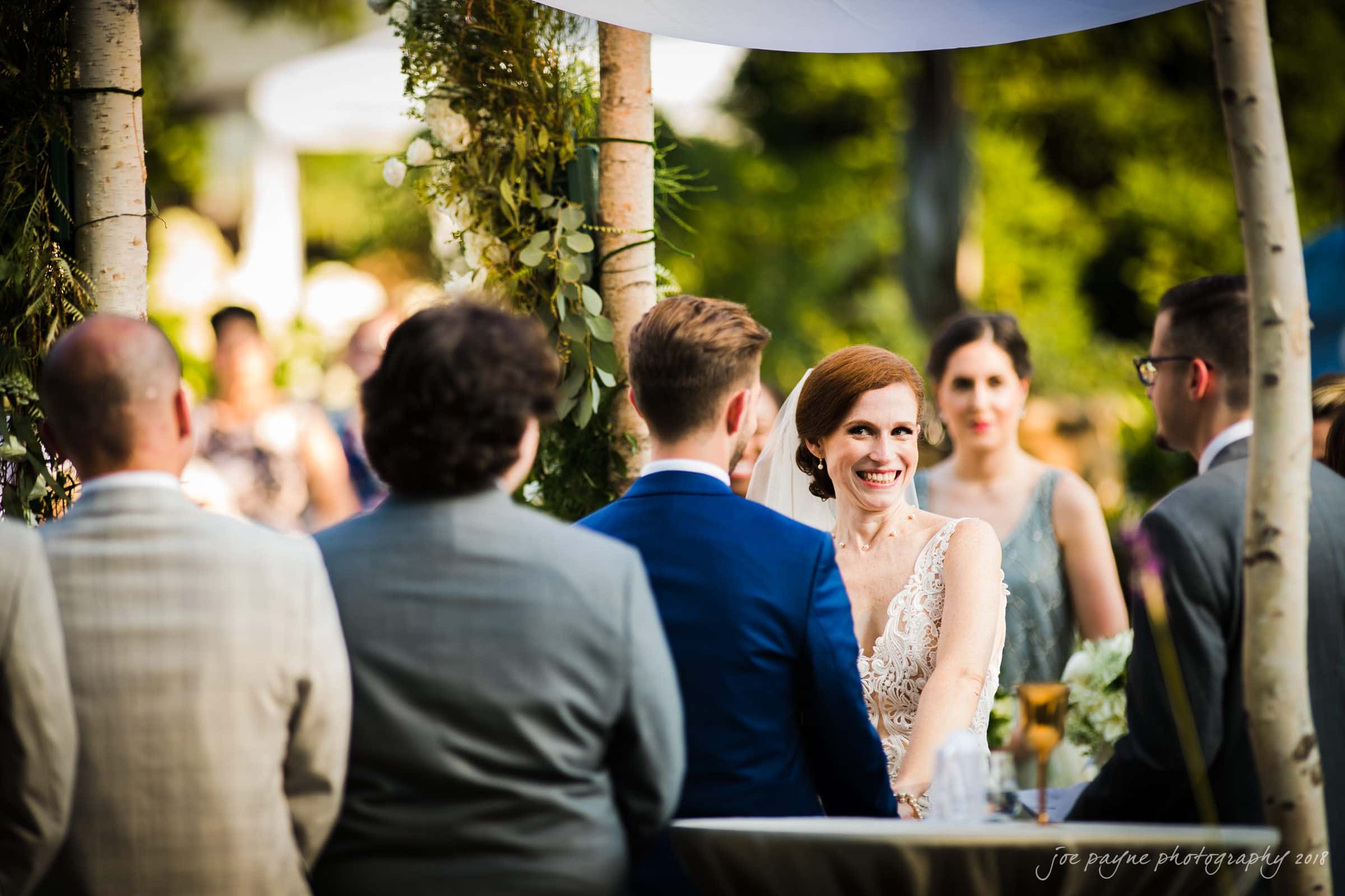 A bride in a white dress smiles at the groom during an outdoor wedding ceremony. They are surrounded by guests under a decorated arch, with greenery and sunlight in the background.
