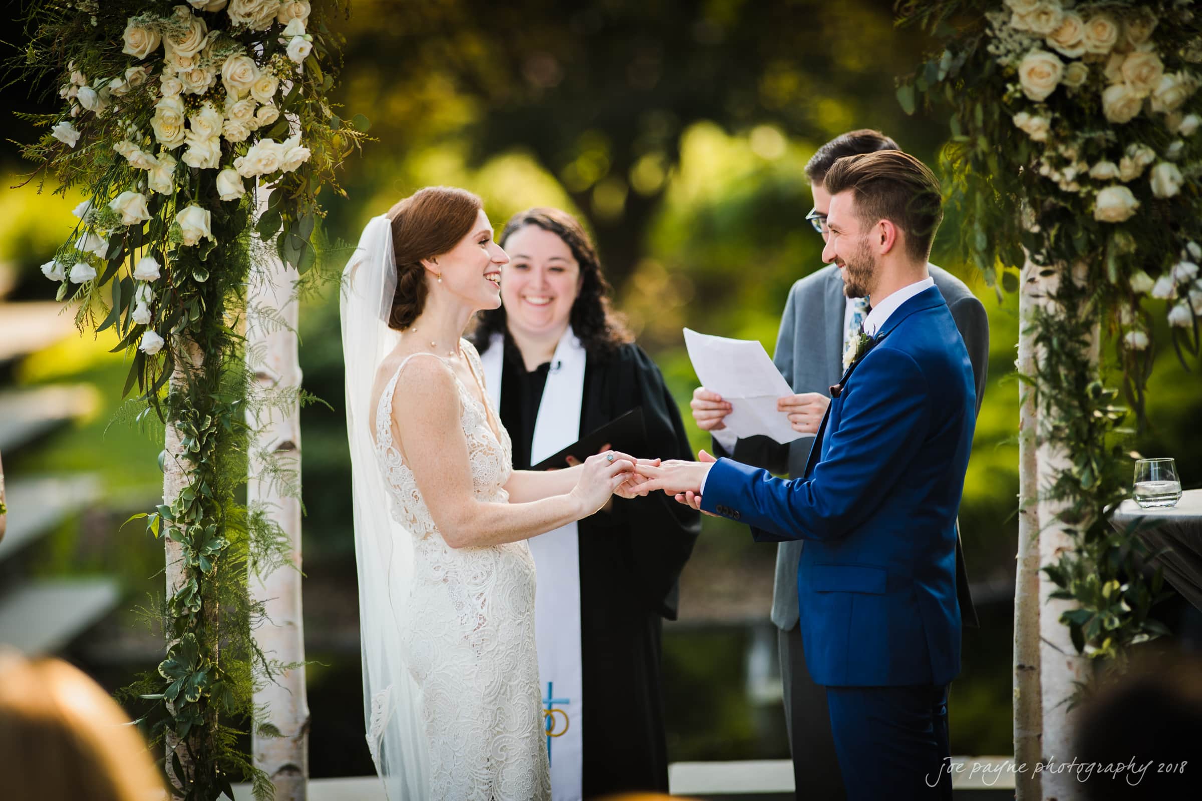 A bride and groom exchange rings under a floral arch during an outdoor wedding ceremony. They are smiling at each other while a person in religious attire officiates. The background is lush and green.