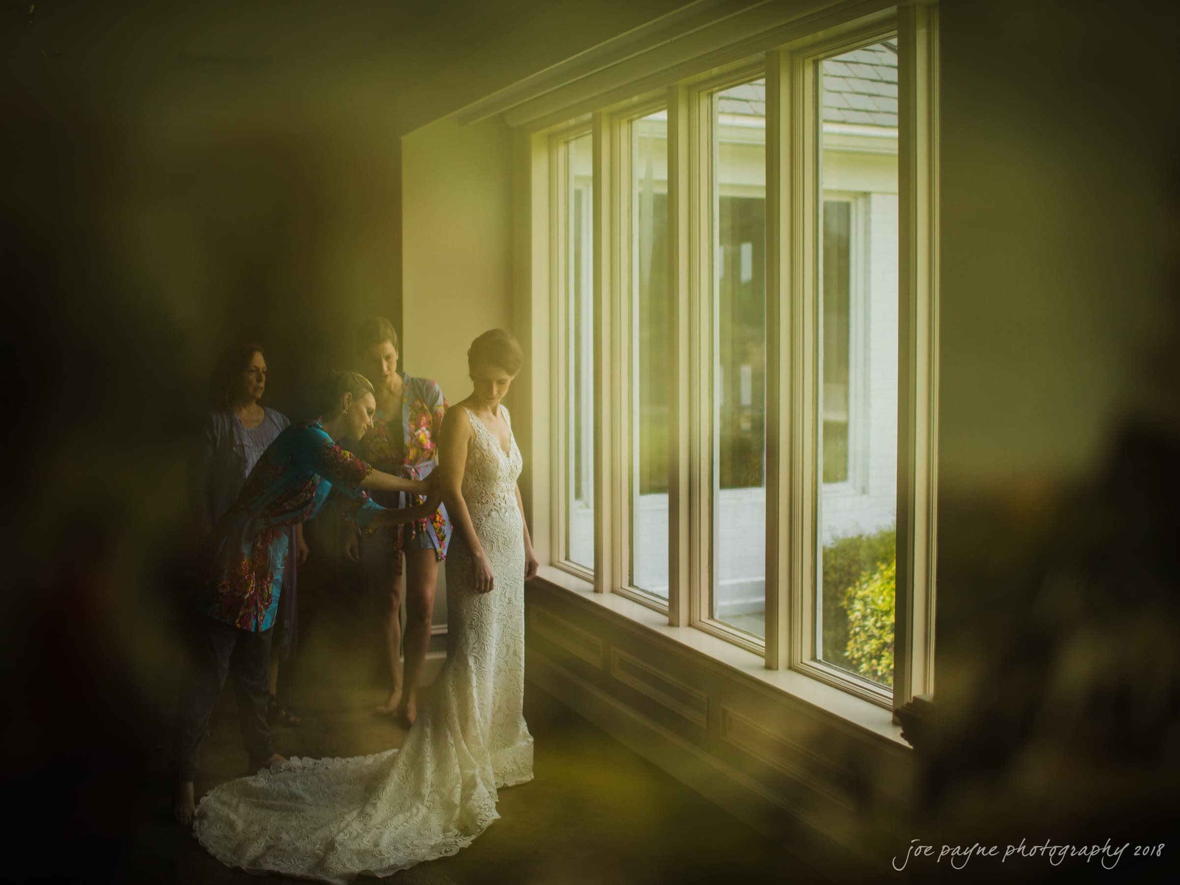 A bride in a lace wedding dress stands by a large window. Several people assist her with her gowns train. Natural light illuminates the room, creating a serene atmosphere.