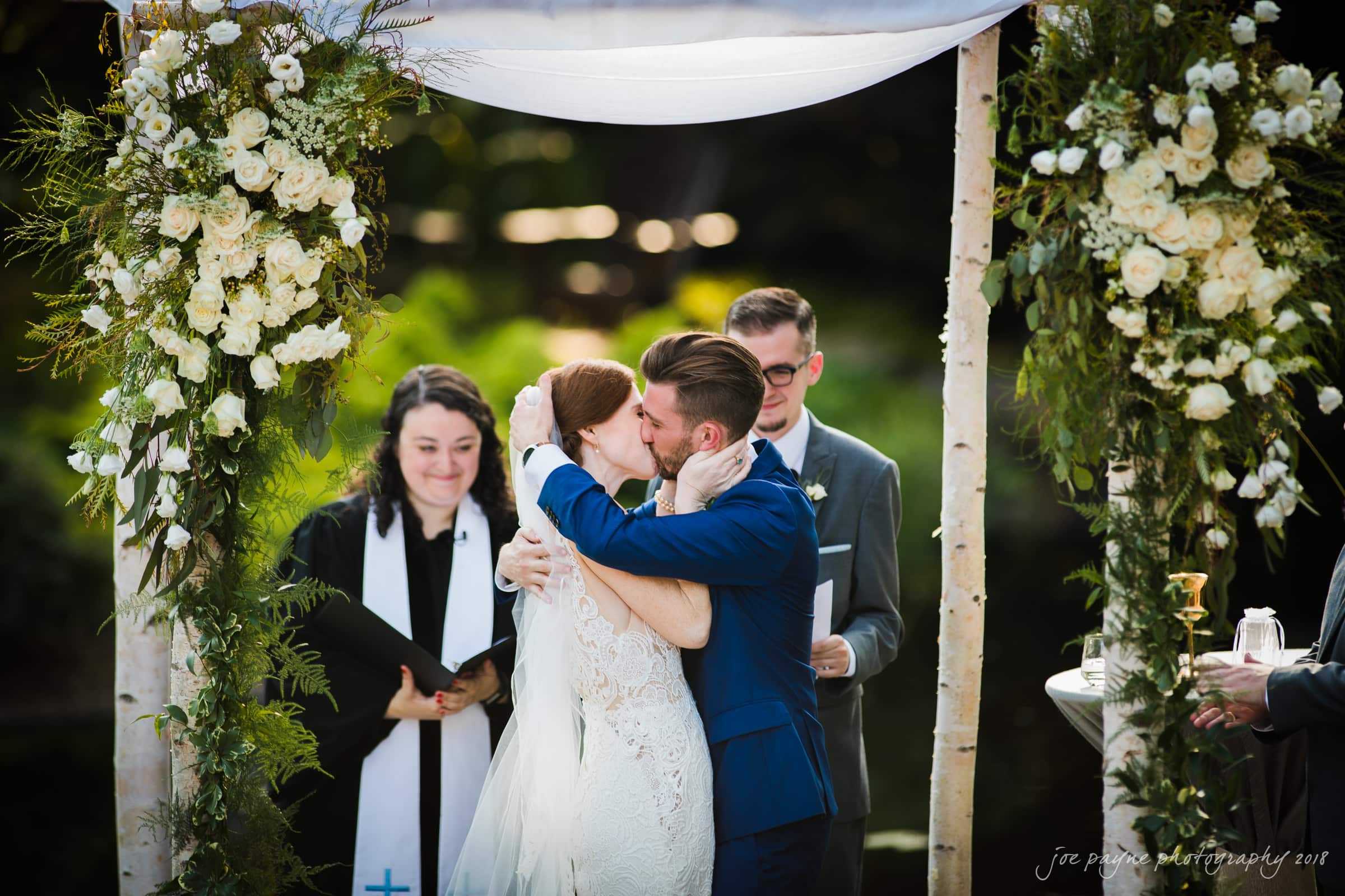 A bride and groom share a kiss under a floral arch during an outdoor wedding ceremony. The officiant and another person stand nearby, smiling. The setting is lush and green, with a sense of celebration and joy.