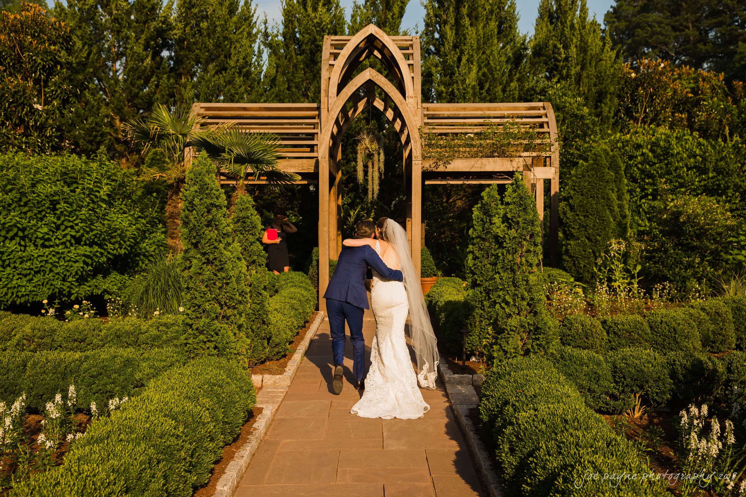 A bride and groom walk arm in arm down a garden path. The groom is in a blue suit, and the bride wears a white dress with a long train. They approach a wooden arch, surrounded by lush greenery under a sunny sky.