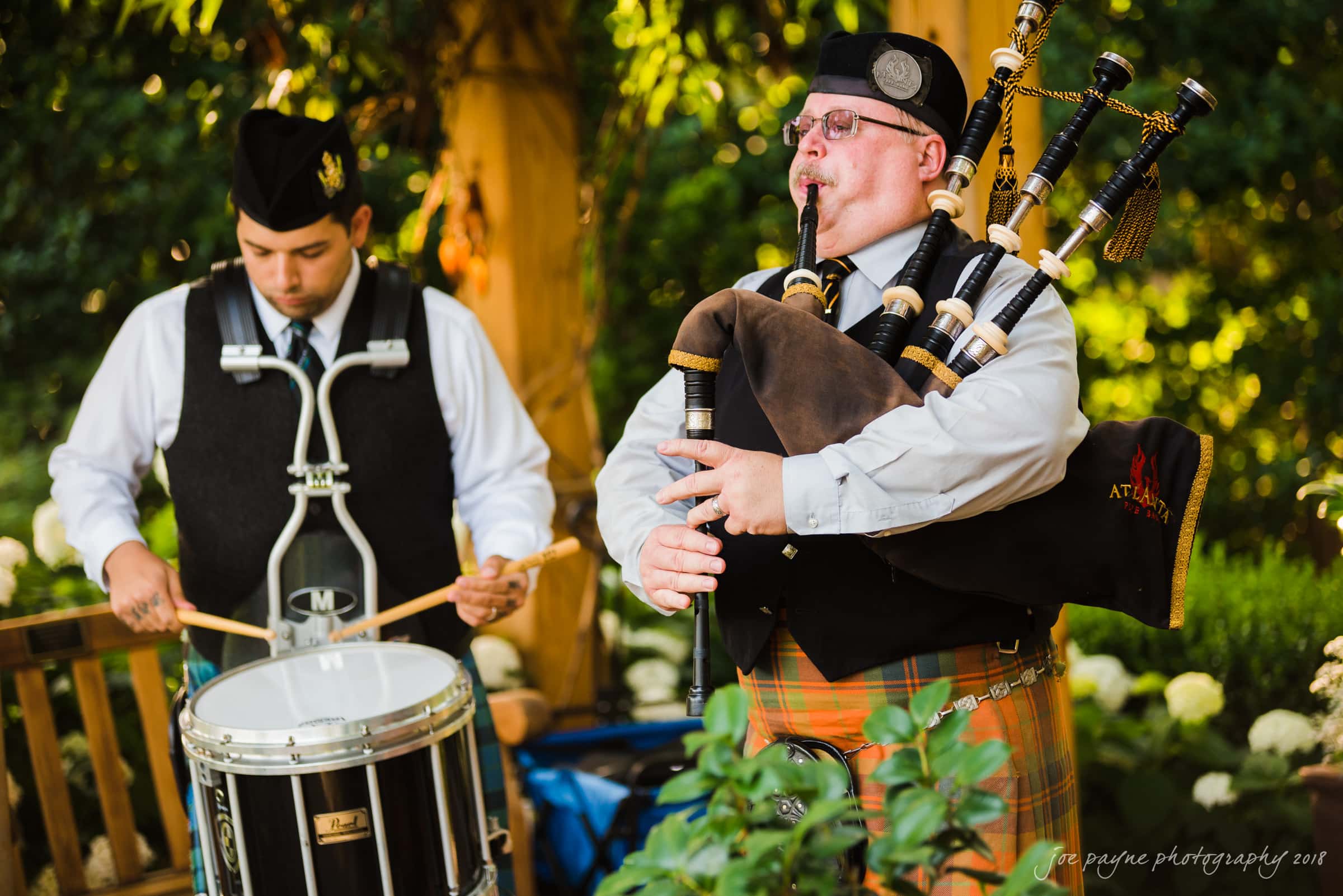 A bagpiper in a kilt plays the bagpipes while a drummer in a uniform plays a snare drum. They are outdoors, surrounded by greenery and flowers, performing on a sunny day.