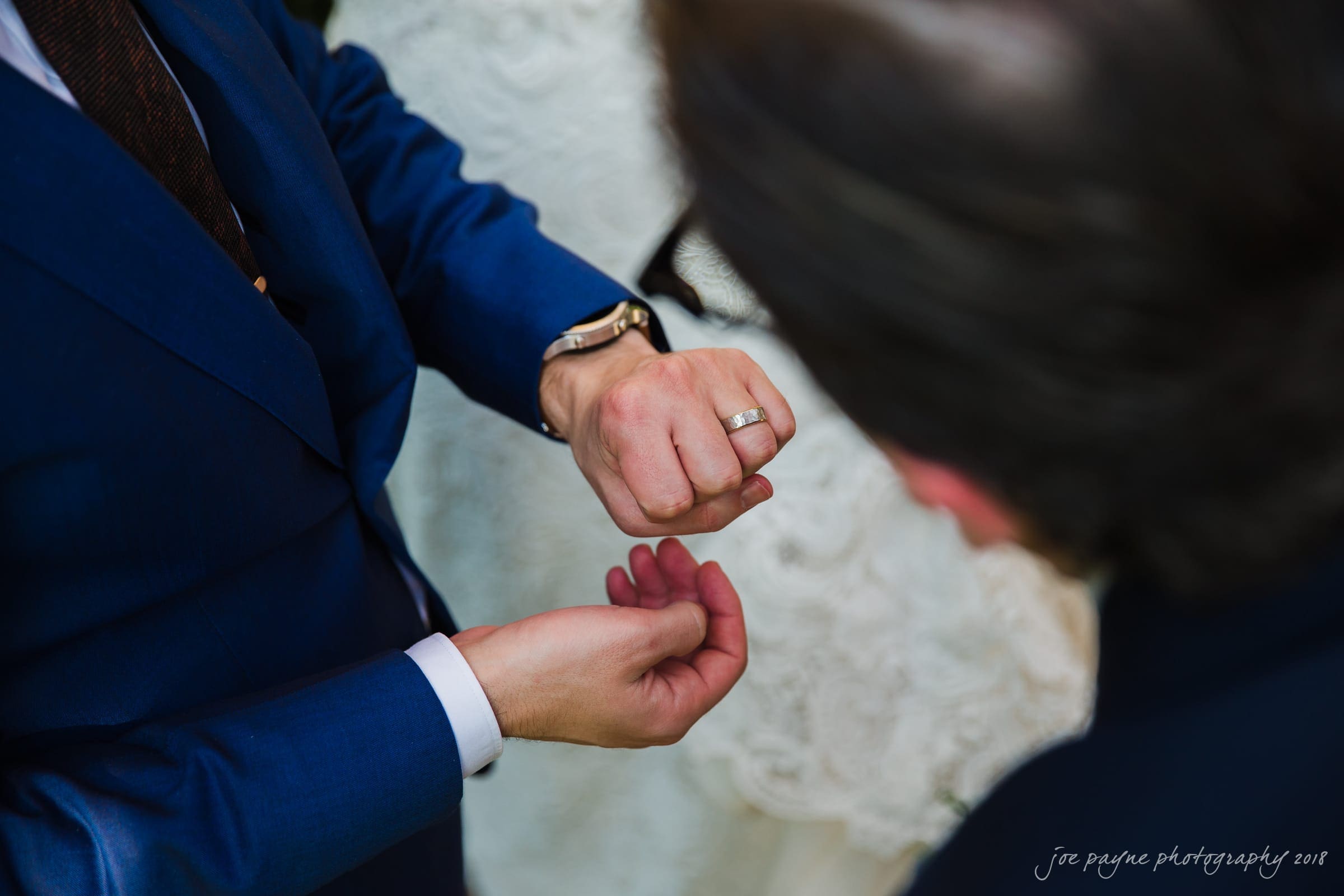 Two people in formal attire are adjusting a wristwatch and shirt cuffs. One wears a wedding ring and a blue suit, while the other appears partially in the foreground. The interaction seems focused and attentive.