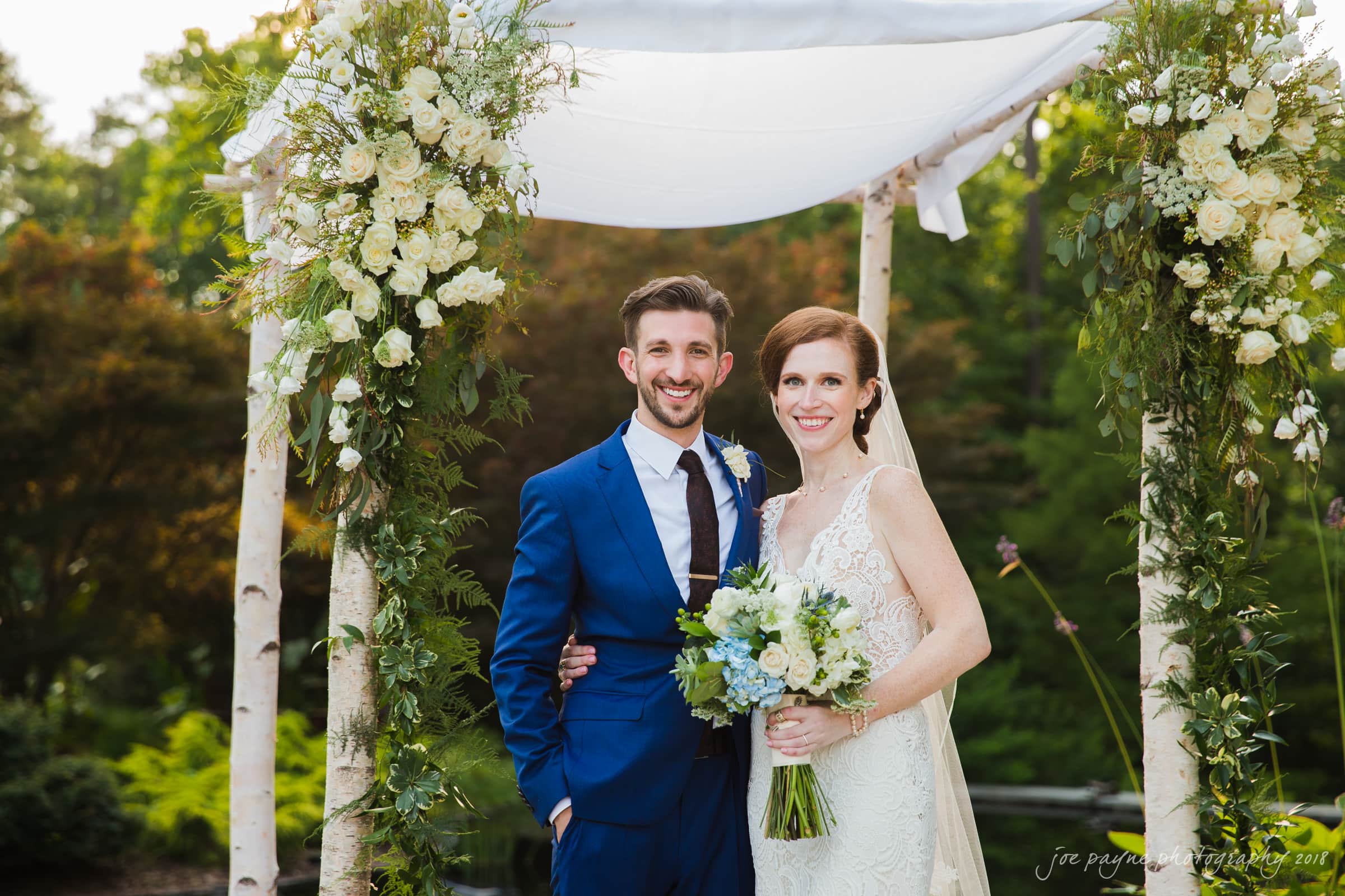 A bride and groom stand smiling under a floral arch. The bride wears a lace dress and holds a bouquet, while the groom wears a blue suit. Greenery and white flowers decorate the arch. The background is filled with lush trees.