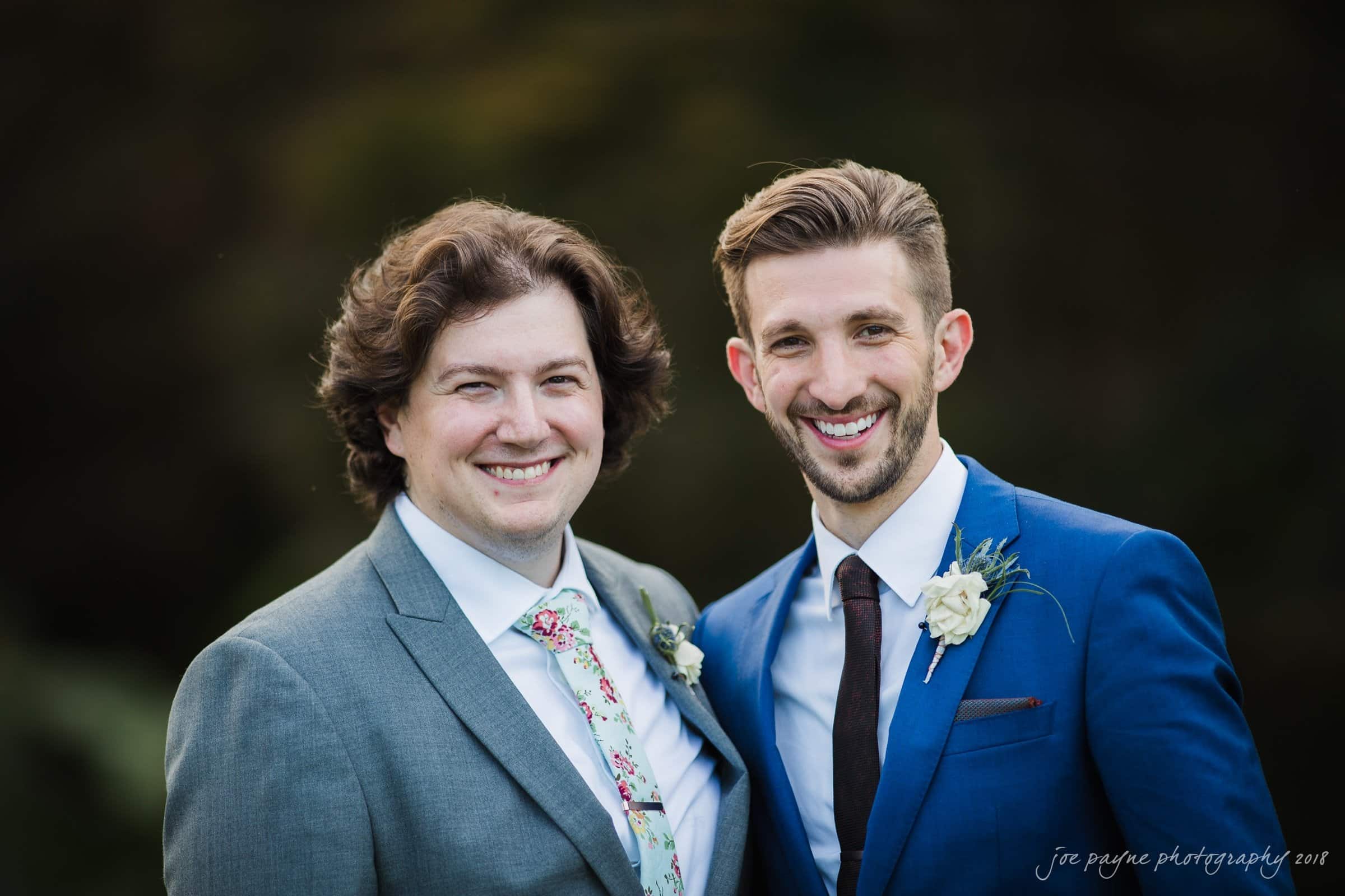 Two smiling people in suits stand outdoors against a dark, blurred background. One wears a gray suit with a floral tie, and the other wears a navy suit with a white shirt and tie. Both have boutonnières on their lapels.