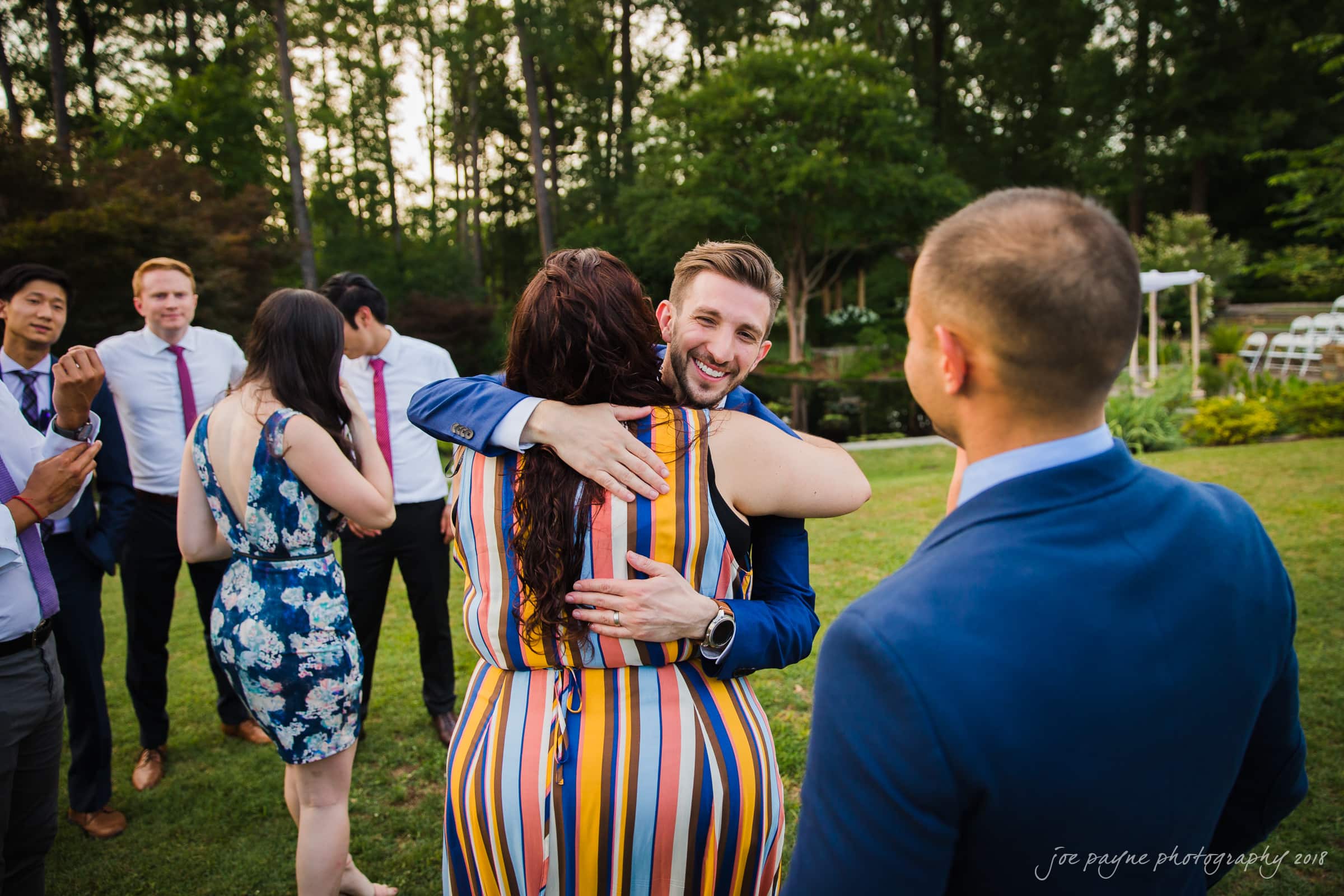 A group of people are gathered outdoors, enjoying a social event. A man in a suit is smiling and hugging a woman wearing a striped dress, while others are chatting in the background amidst lush greenery.