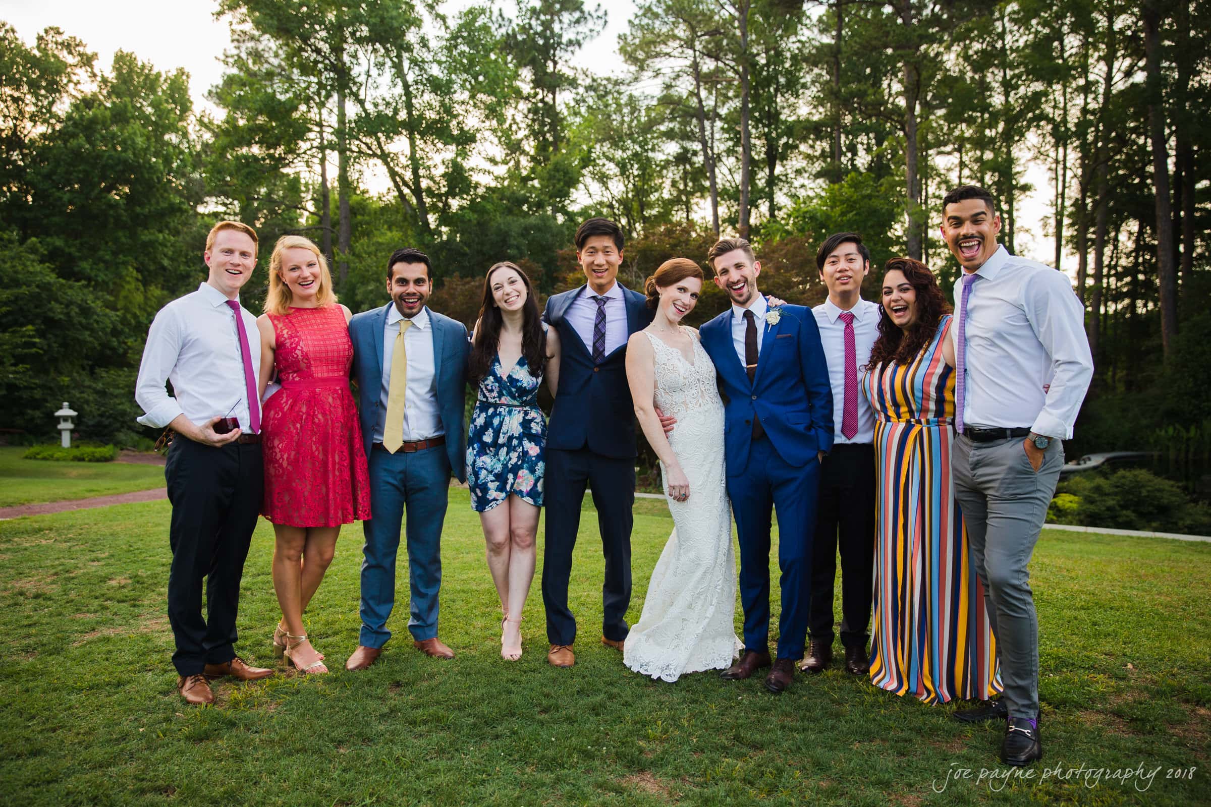 A group of ten people, dressed in formal and semi-formal attire, stand closely together outdoors on a grassy area with trees in the background. The group is smiling and posing for the photo in a relaxed and cheerful manner.