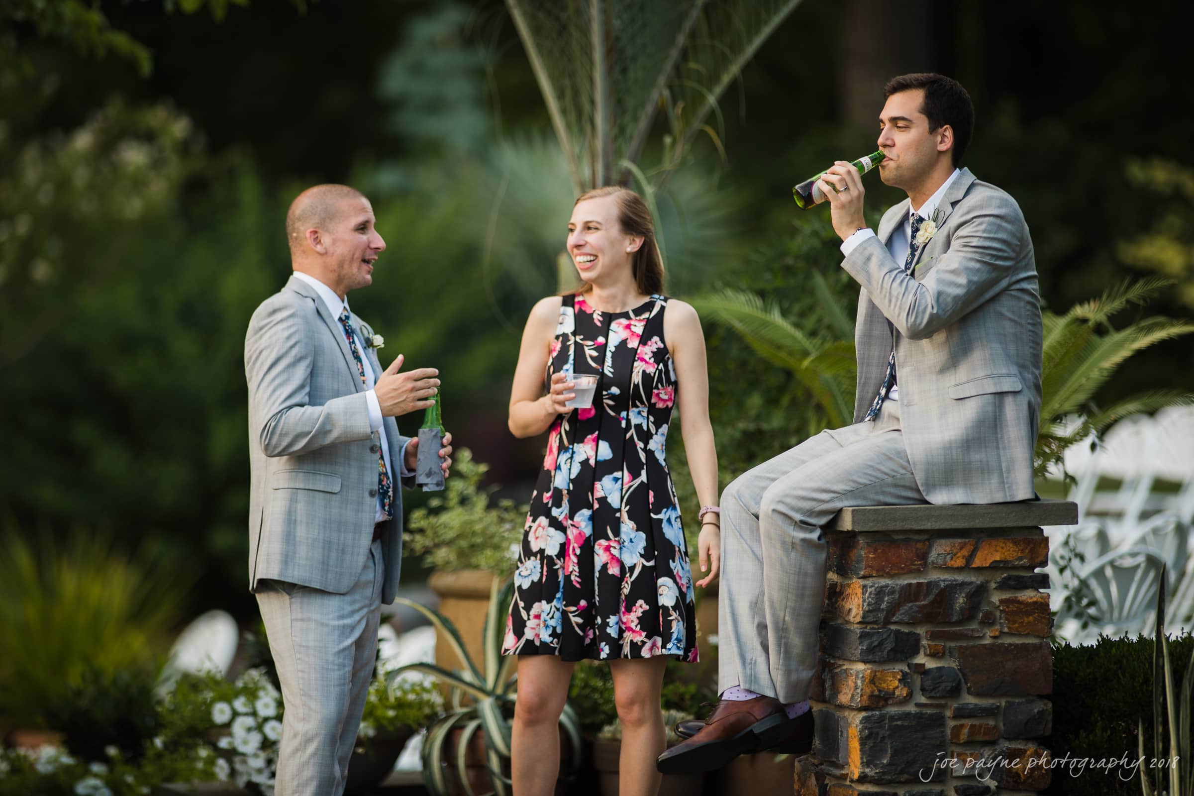Three people in formal attire are enjoying drinks outdoors. Two men in gray suits, one standing and one sitting on a stone ledge, chat with a woman in a floral dress. They are surrounded by greenery.