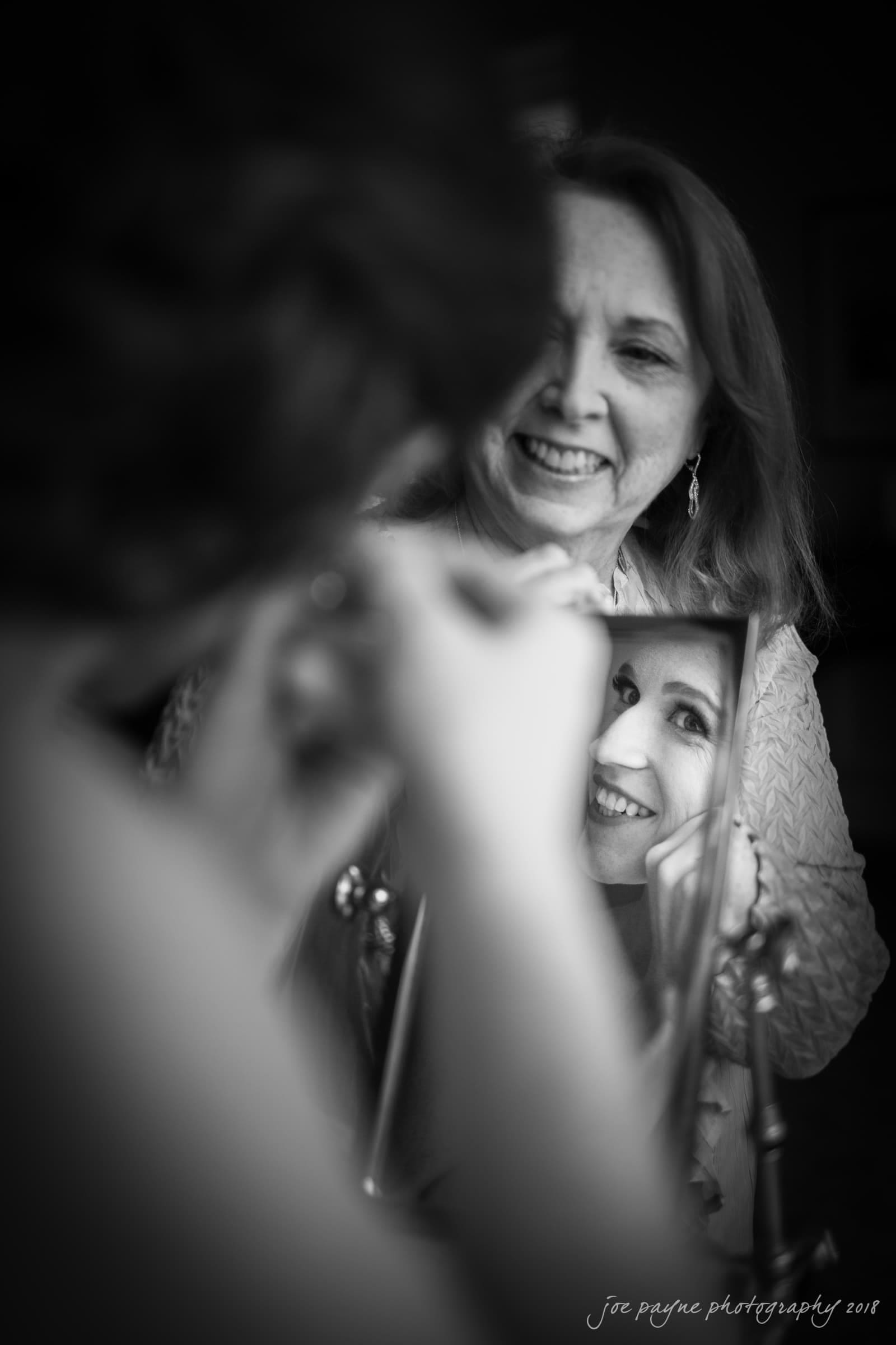 A woman is smiling as she holds a mirror reflecting the face of another woman who is applying makeup. The image is in black and white, capturing an intimate and joyful moment.
