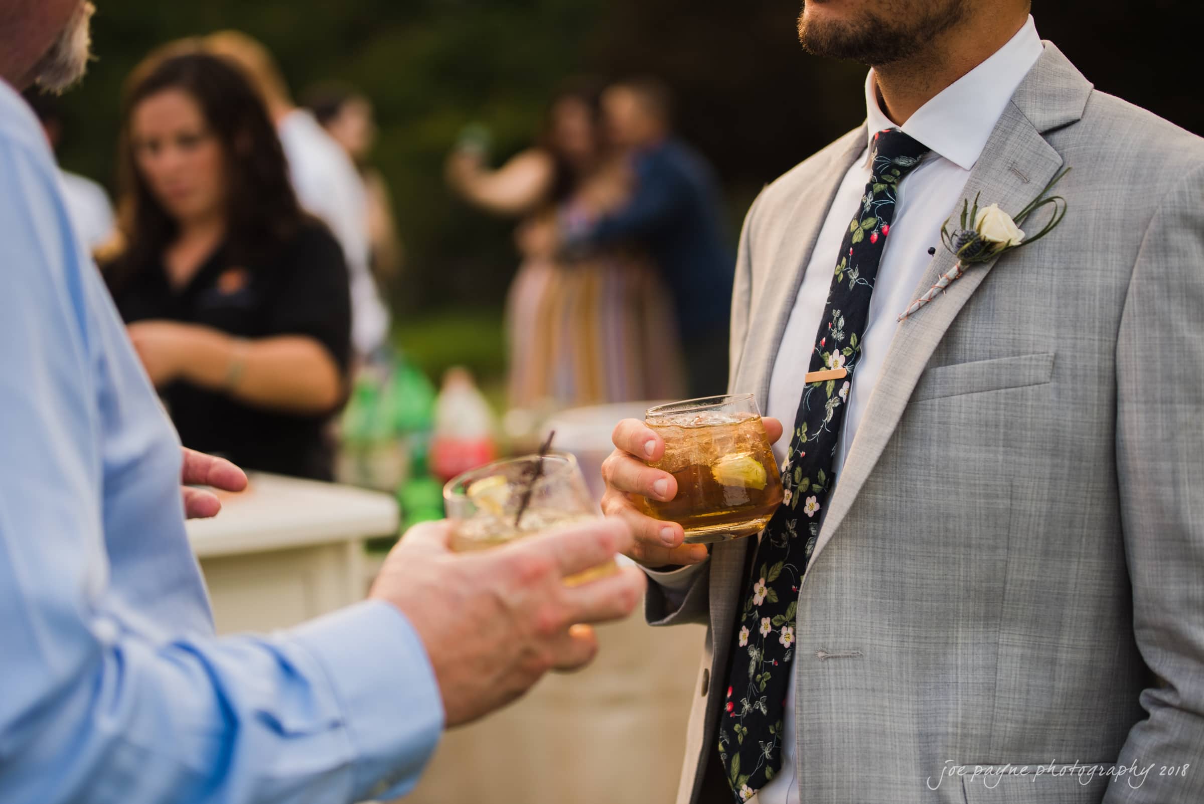 Two men in suits holding drinks engage in conversation outdoors. One wears a light gray suit with a floral tie and boutonniere. Blurred people and greenery are visible in the background.