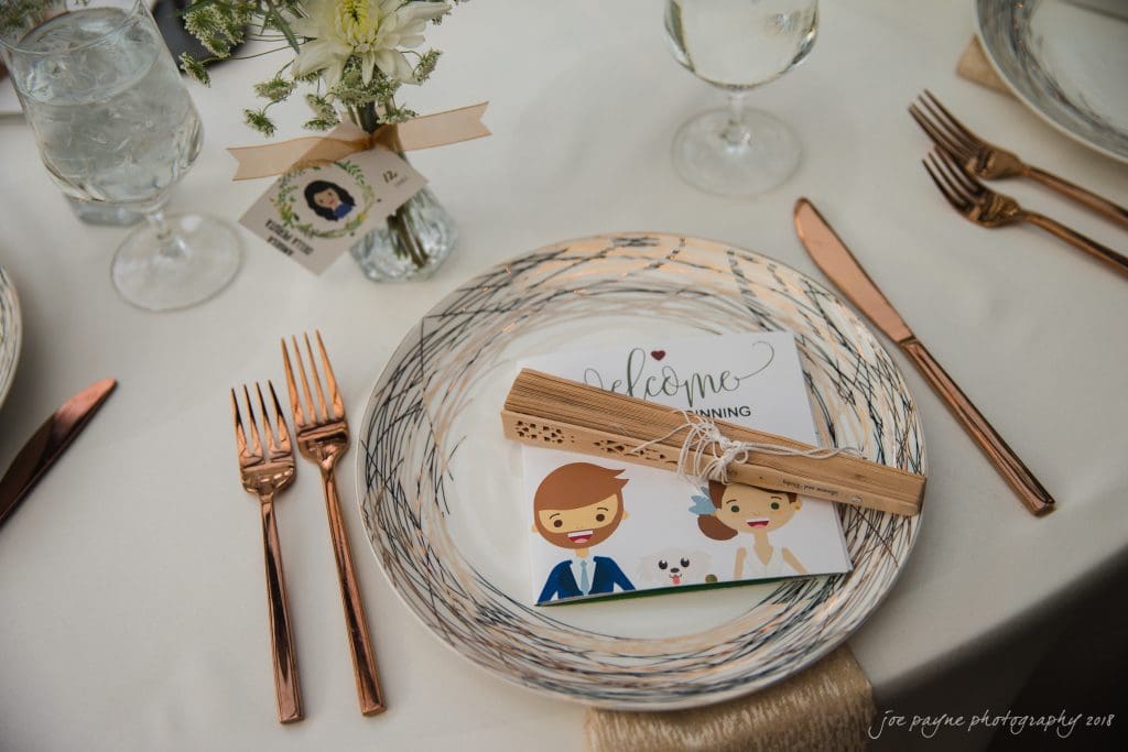 A table setting with a decorative plate, napkin, and menu card featuring cartoon images of a couple. Silverware includes rose gold forks and knives. A small flower arrangement in a vase is beside a wine glass and a water glass with ice.