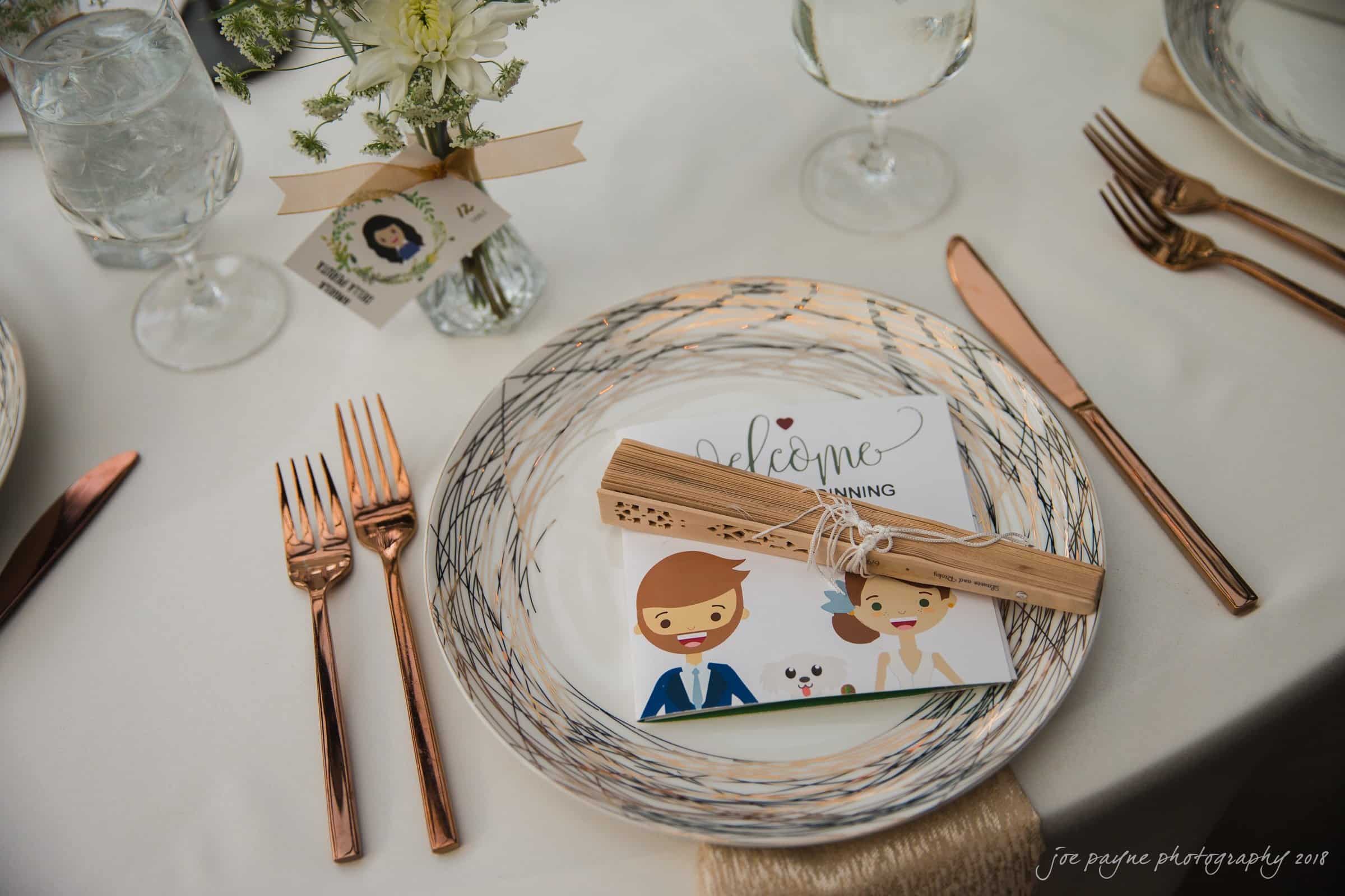 A table setting with a decorative plate, napkin, and menu card featuring cartoon images of a couple. Silverware includes rose gold forks and knives. A small flower arrangement in a vase is beside a wine glass and a water glass with ice.