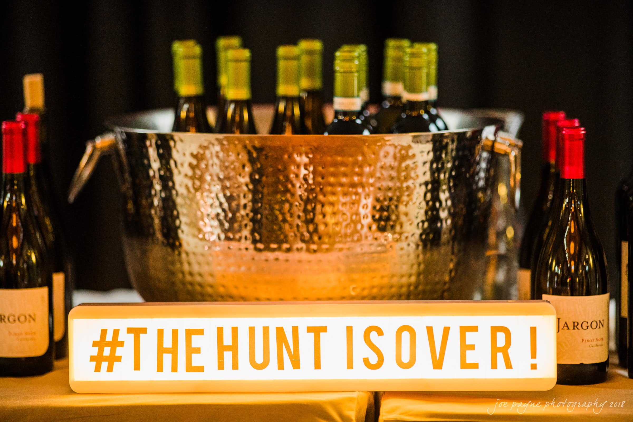 A large metal ice bucket filled with wine bottles sits on a table. In front of it is a lit sign displaying the text #THE HUNT IS OVER! Bottles are also placed on the table around the bucket.