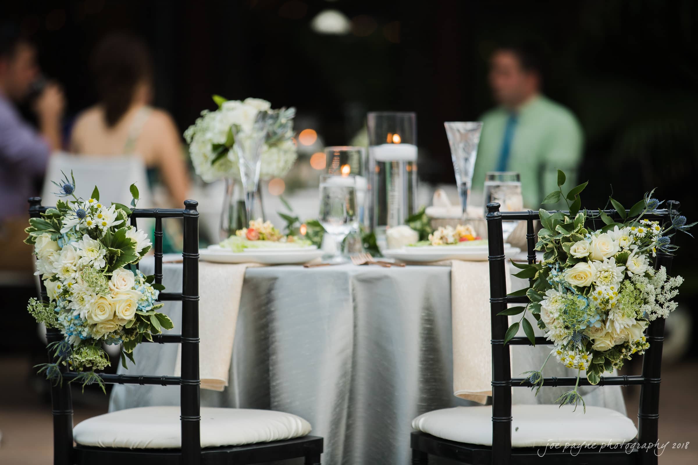 An elegantly set table with a light gray tablecloth, adorned with white floral arrangements and candles. Two black chairs with floral decorations face the table. Blurred figures are in the background, suggesting a formal event.