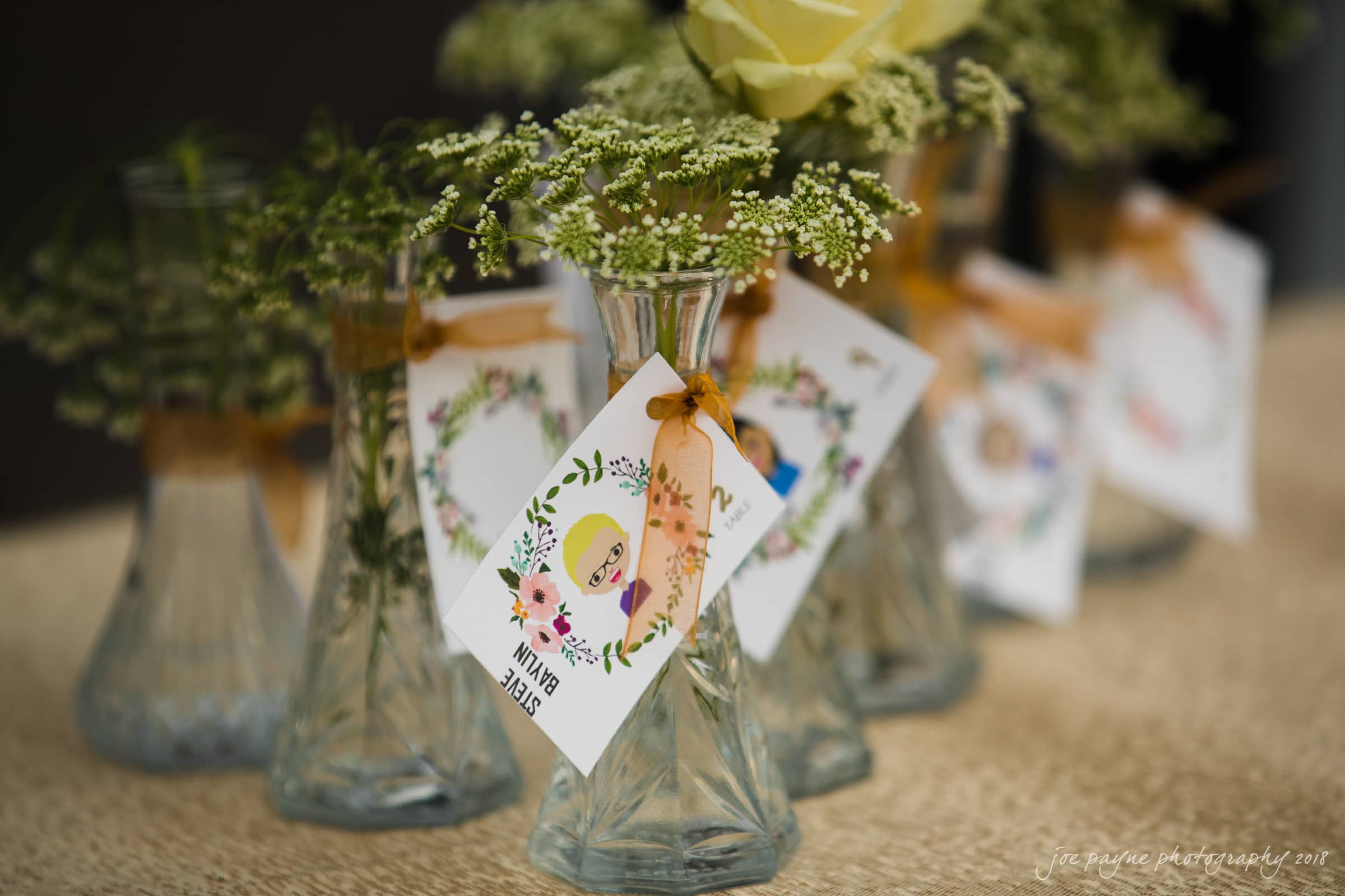 Close-up of small glass vases with floral decorations and personalized tags tied with ribbon. The vases contain delicate green leaves and a single yellow rose. Tags feature colorful illustrations and names, on a textured beige surface.