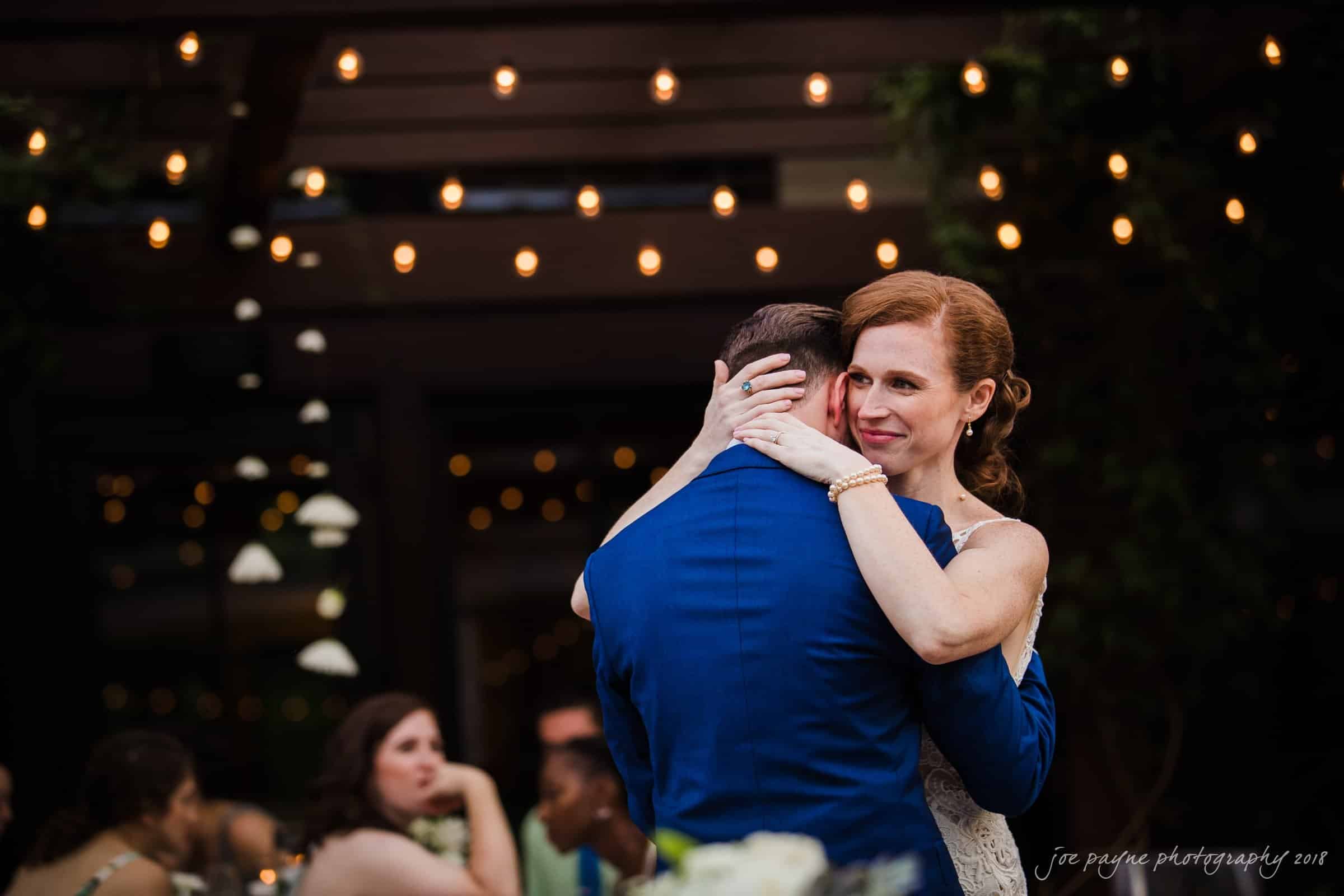 A bride and groom share a dance under string lights. The bride, in a white dress, embraces the groom in a blue suit, smiling softly. Guests are seated in the background.