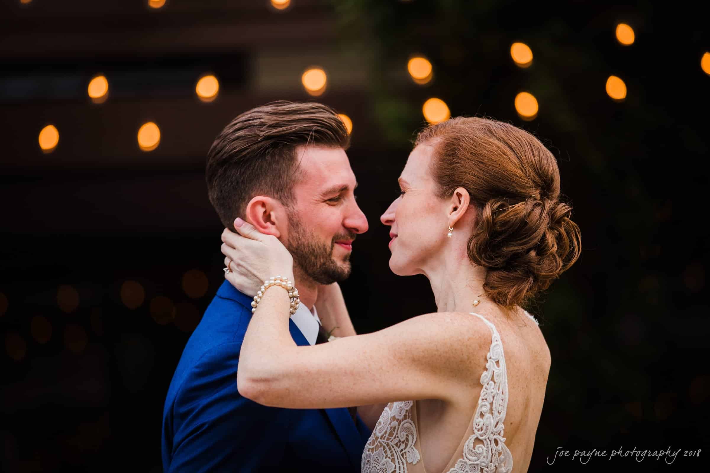 A groom in a blue suit and a bride in a lace dress embrace and smile at each other under warm string lights.