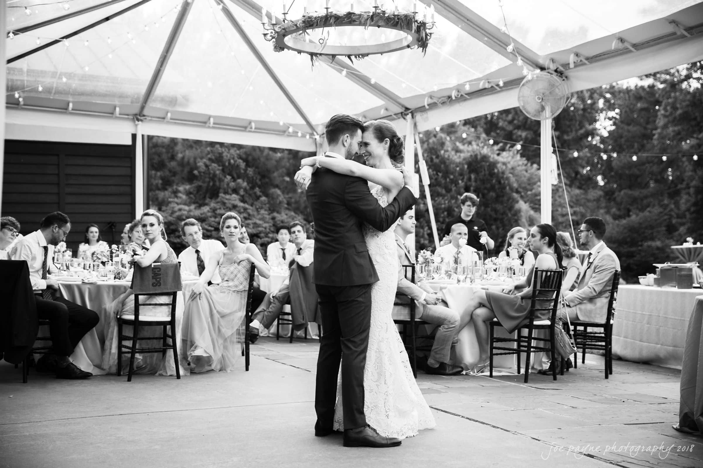 A bride and groom share their first dance under a clear tent with an elegant chandelier. Guests sit at round tables, watching the couple. The setting has a romantic and intimate atmosphere.