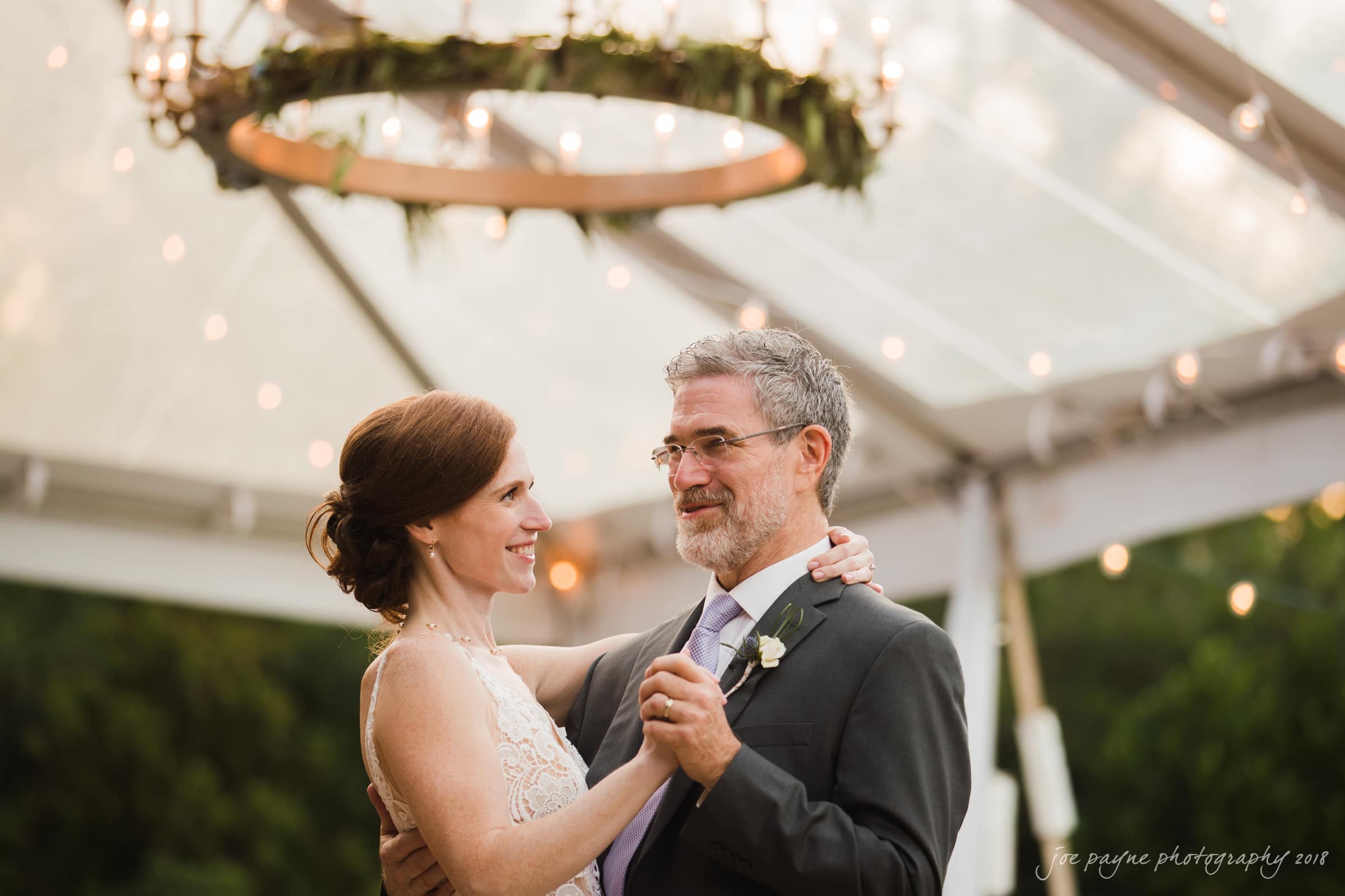 A couple dances under a decorative circular chandelier with greenery at an outdoor event. The woman is wearing a white dress, and the man is in a gray suit. They are smiling at each other, surrounded by soft lighting and a forest backdrop.