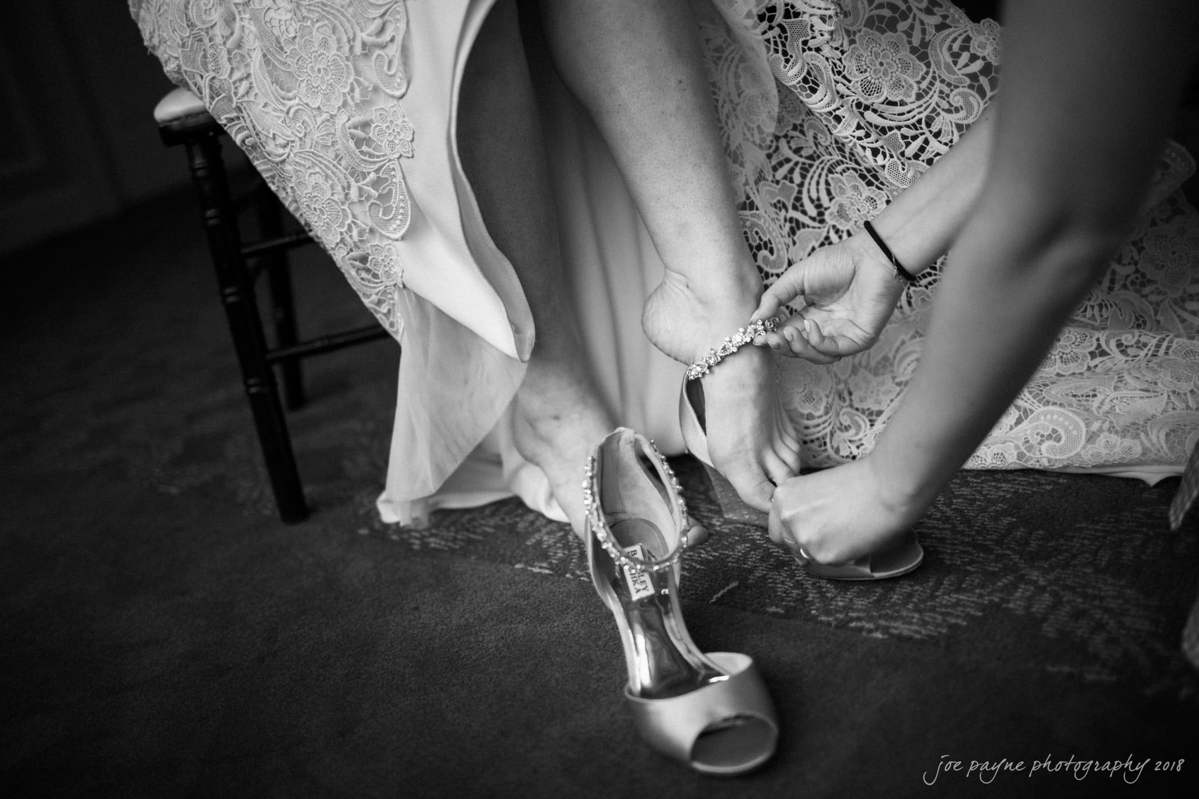 A black-and-white image of a bride sitting, lifting her lace dress slightly as someone helps fasten a delicate anklet on her foot. One high-heeled shoe is on, while the other rests on the floor nearby.