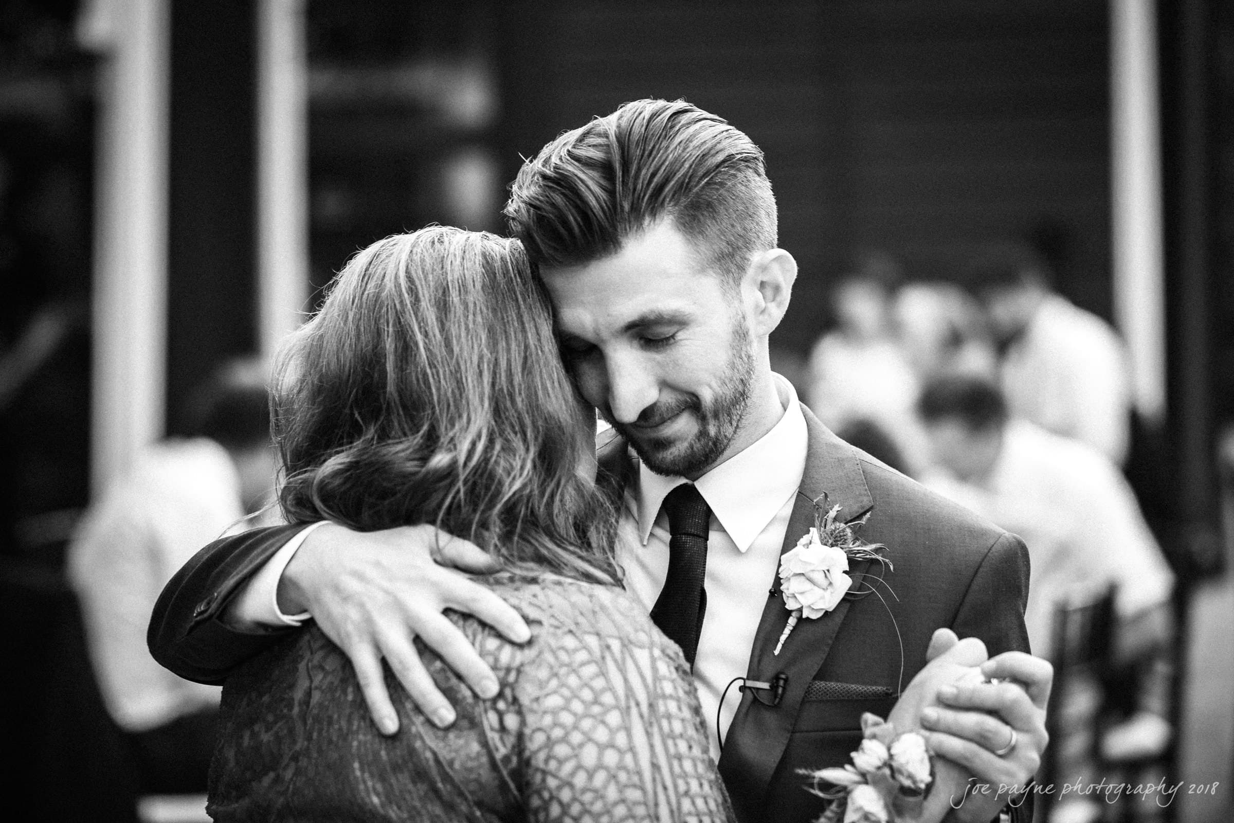 A black-and-white photo of a man in a suit and tie dancing closely with a woman in a patterned dress at an event. The man has a boutonniere on his lapel, and they appear happy and content, surrounded by a blurred background.