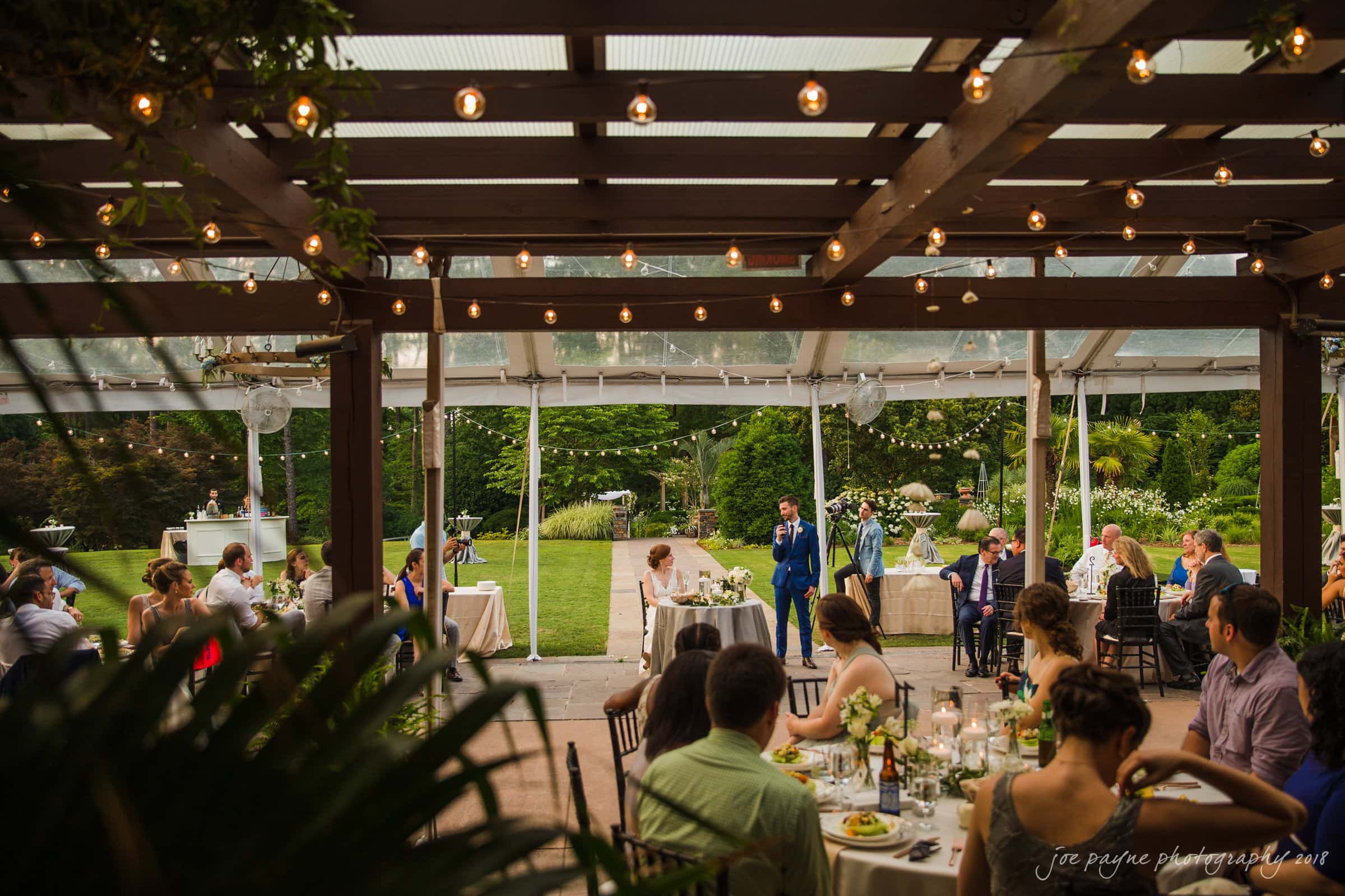 A wedding reception set outdoors under a wooden pergola with string lights. Guests are seated at round tables, while a person in a blue suit gives a speech. Trees and greenery are visible in the background.