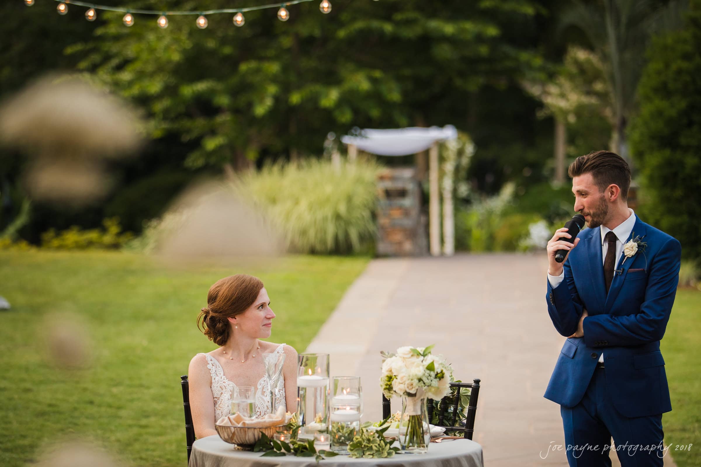 A groom in a blue suit gives a speech with a microphone while standing next to a seated bride in a white dress at an outdoor wedding reception. They are surrounded by greenery and a table with floral decorations and candles.
