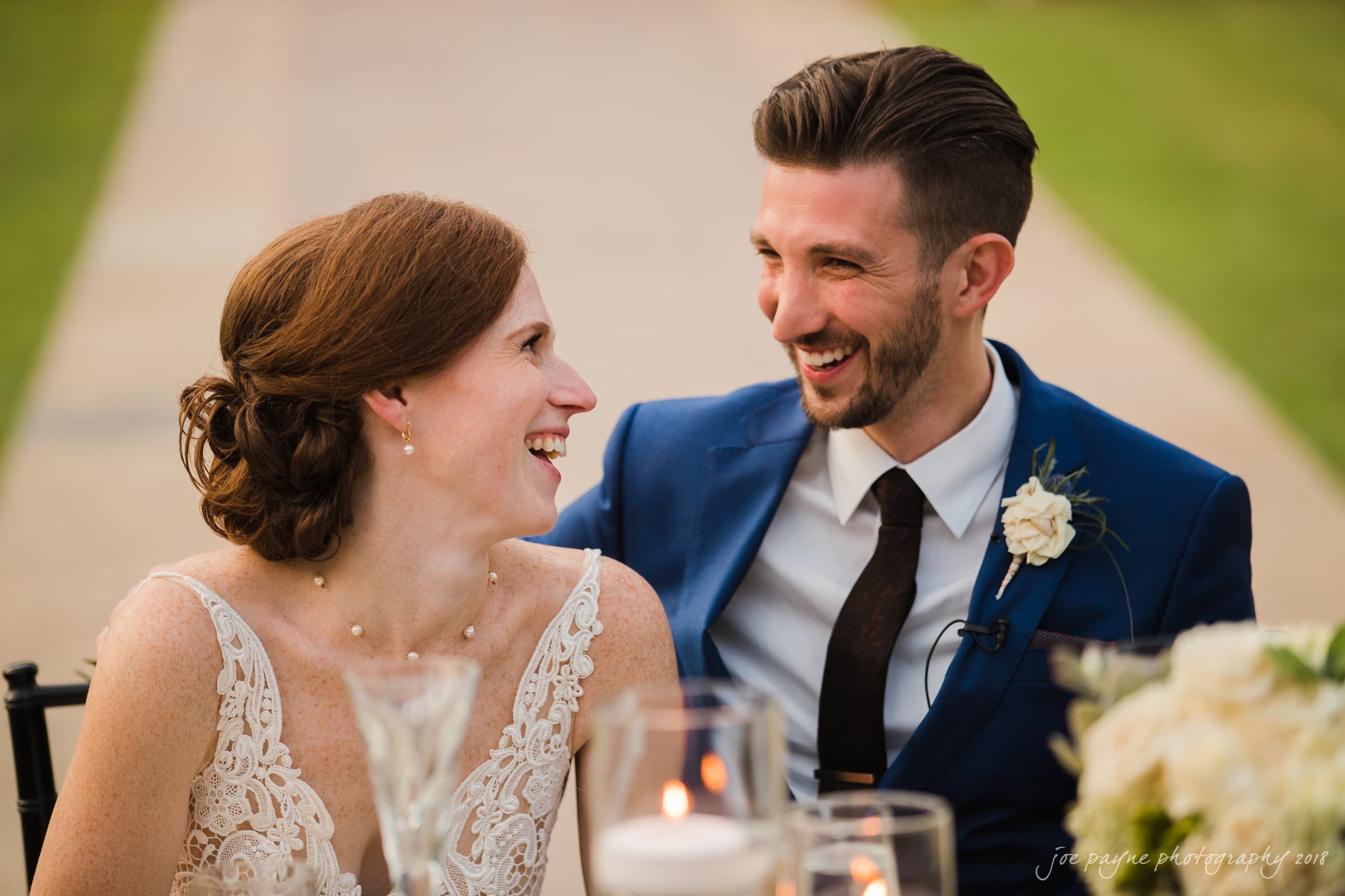 A bride in a white lace dress and a groom in a blue suit share a joyful moment, smiling at each other. They sit at a table with candle decorations and greenery in the background.