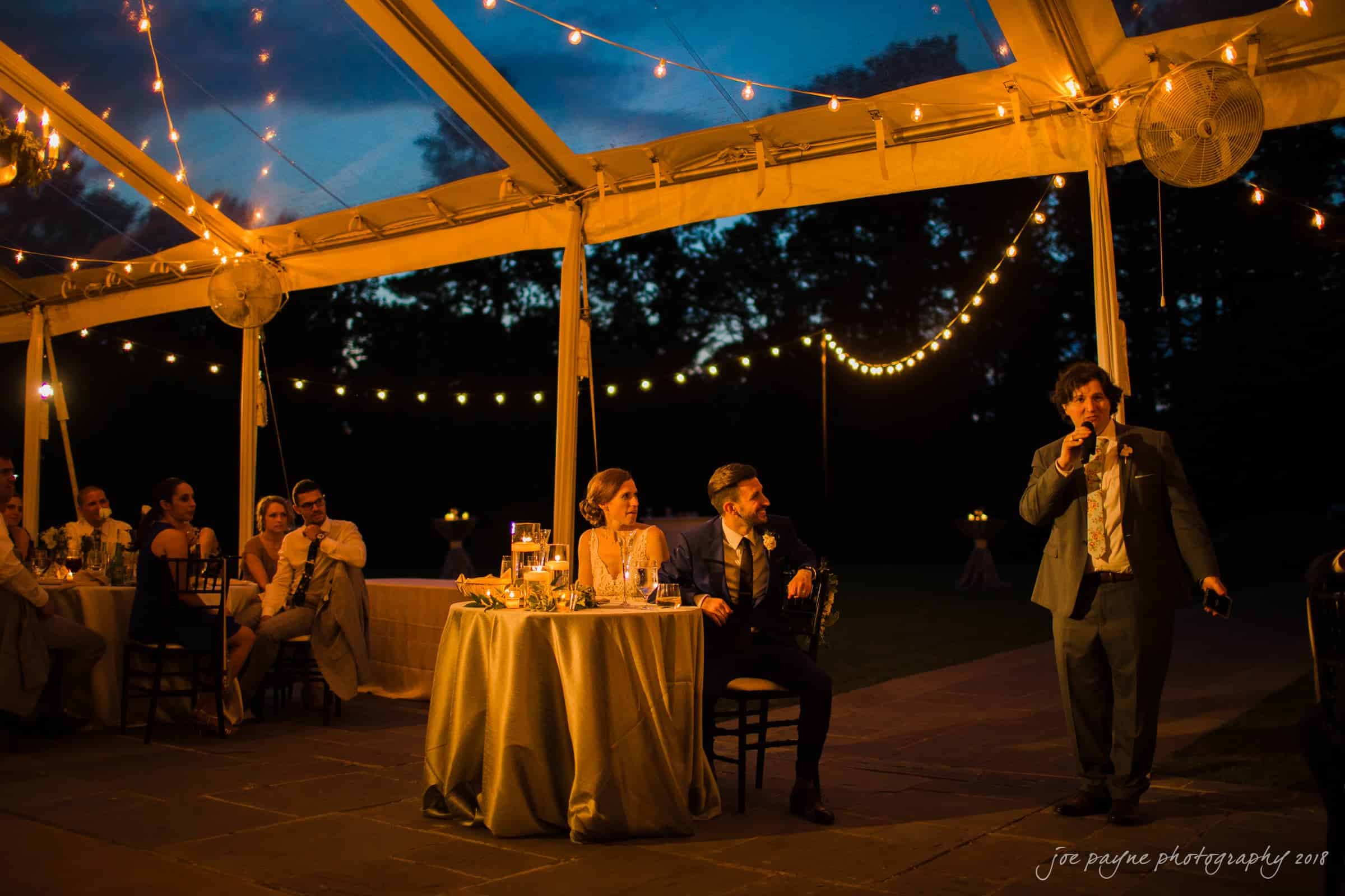 A wedding reception under a tent at night, adorned with string lights. A man in a suit is giving a speech with a microphone. Guests sit at tables, attentively listening. The atmosphere is warm and festive.