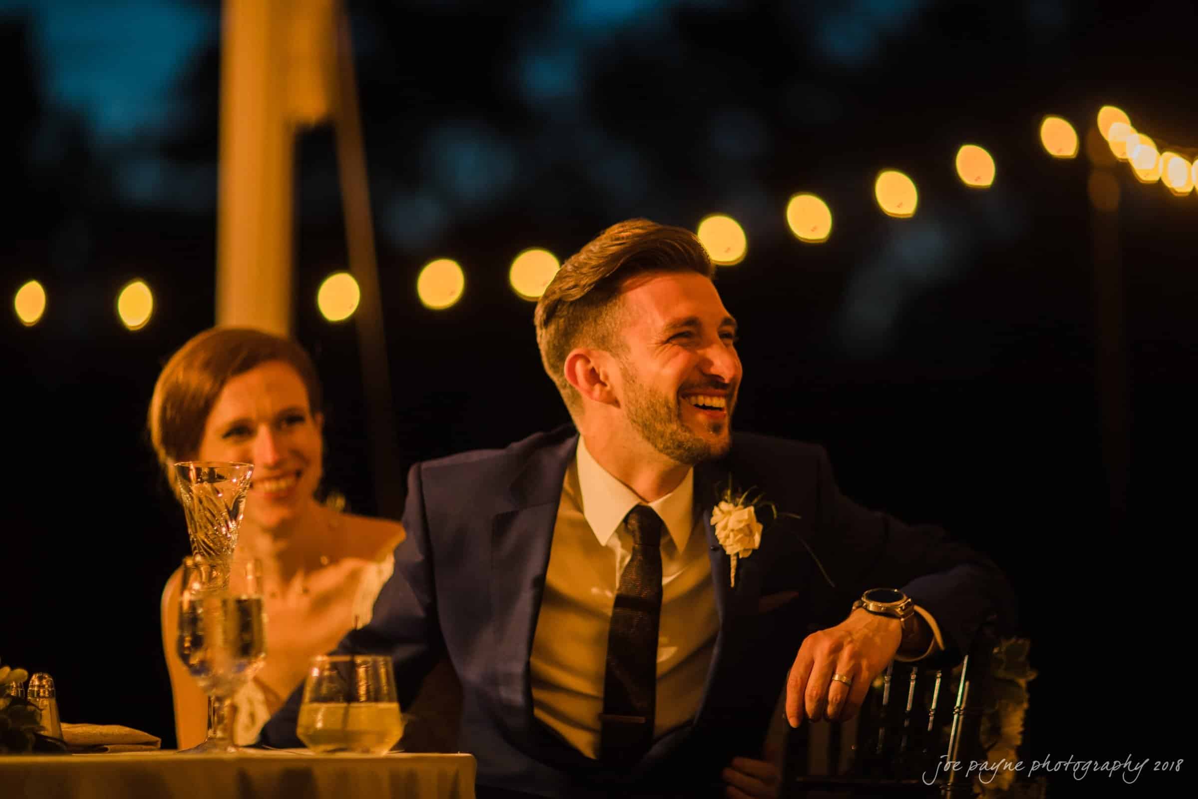 A man in a suit and a woman in a dress sit at a table, smiling and enjoying an evening event. Warm string lights create a cozy, festive atmosphere in the background.