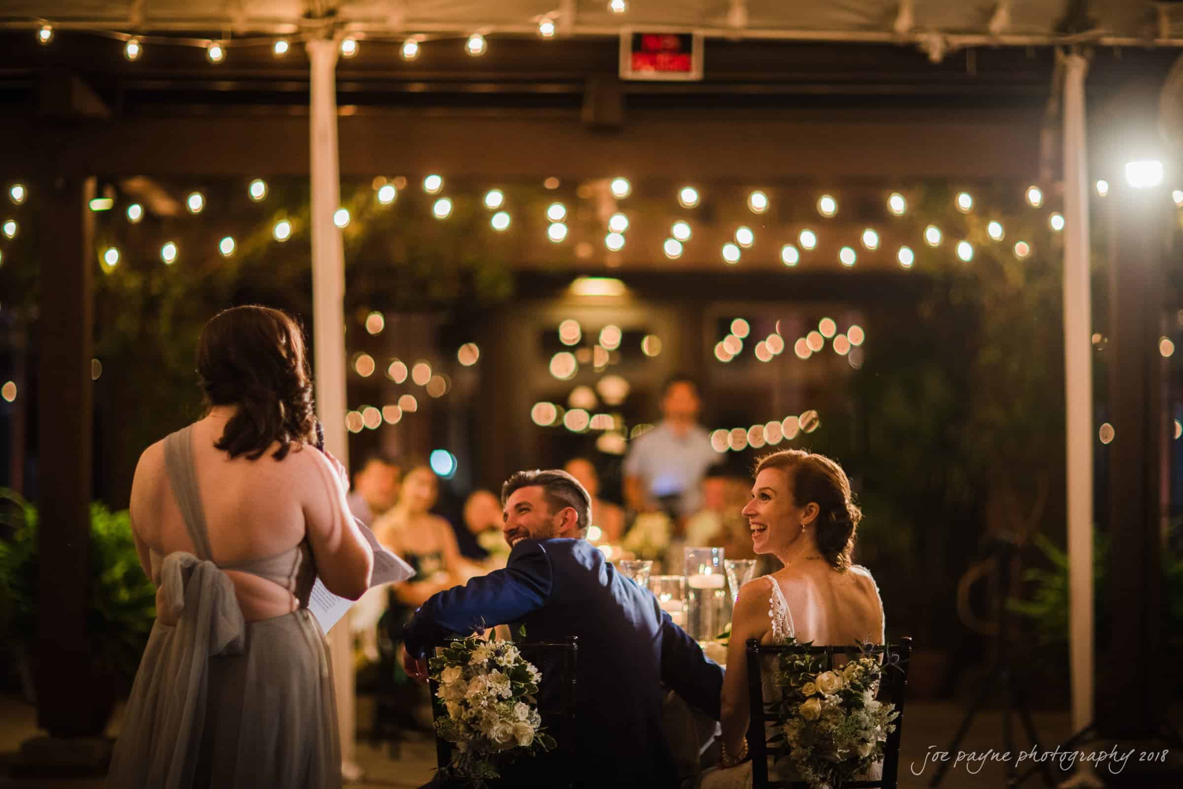 A bride and groom seated at a table outdoors, smiling and listening to a speech. The area is lit with string lights, and guests are visible in the background. Floral arrangements decorate the table.