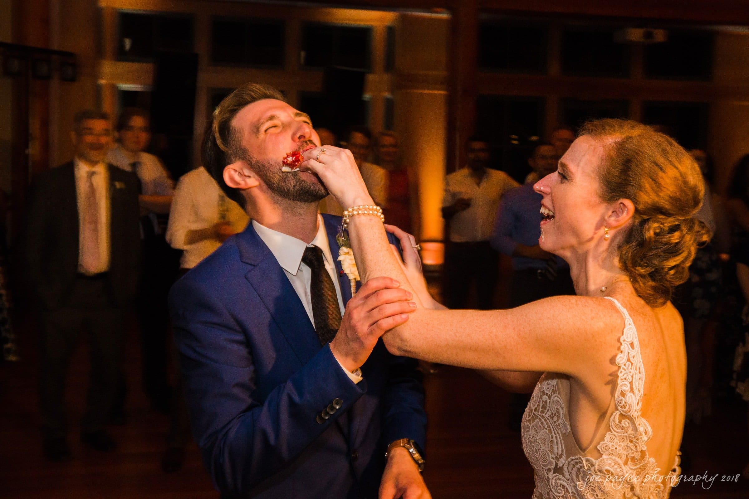 A bride playfully feeds cake to the groom at their wedding reception. Hes laughing, wearing a blue suit, while she is smiling in a white lace dress. Guests are watching in the softly lit background.