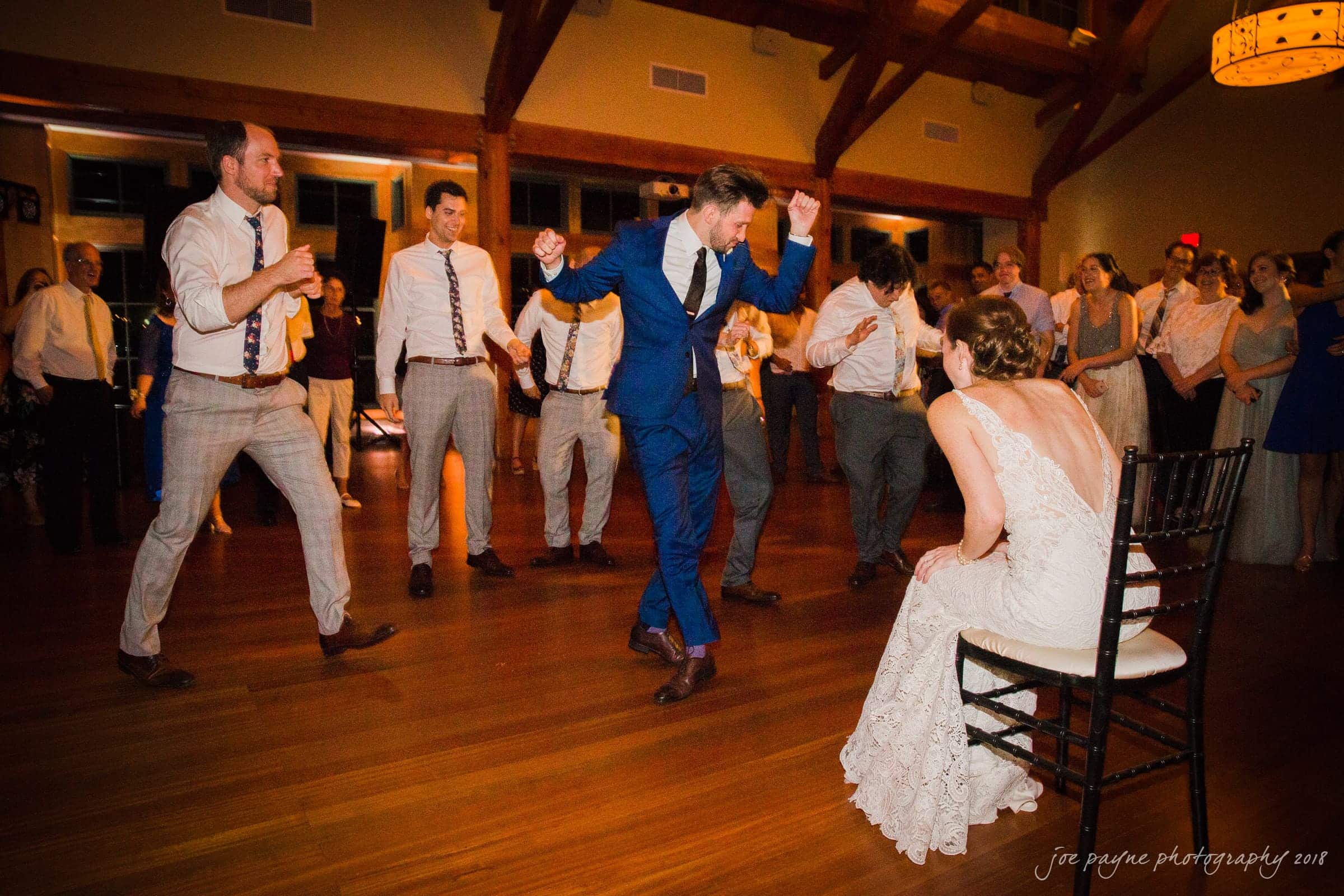 A groom in a blue suit dances enthusiastically in front of a bride seated on a chair, wearing a white lace dress. They are surrounded by groomsmen and guests in a warmly lit reception hall.