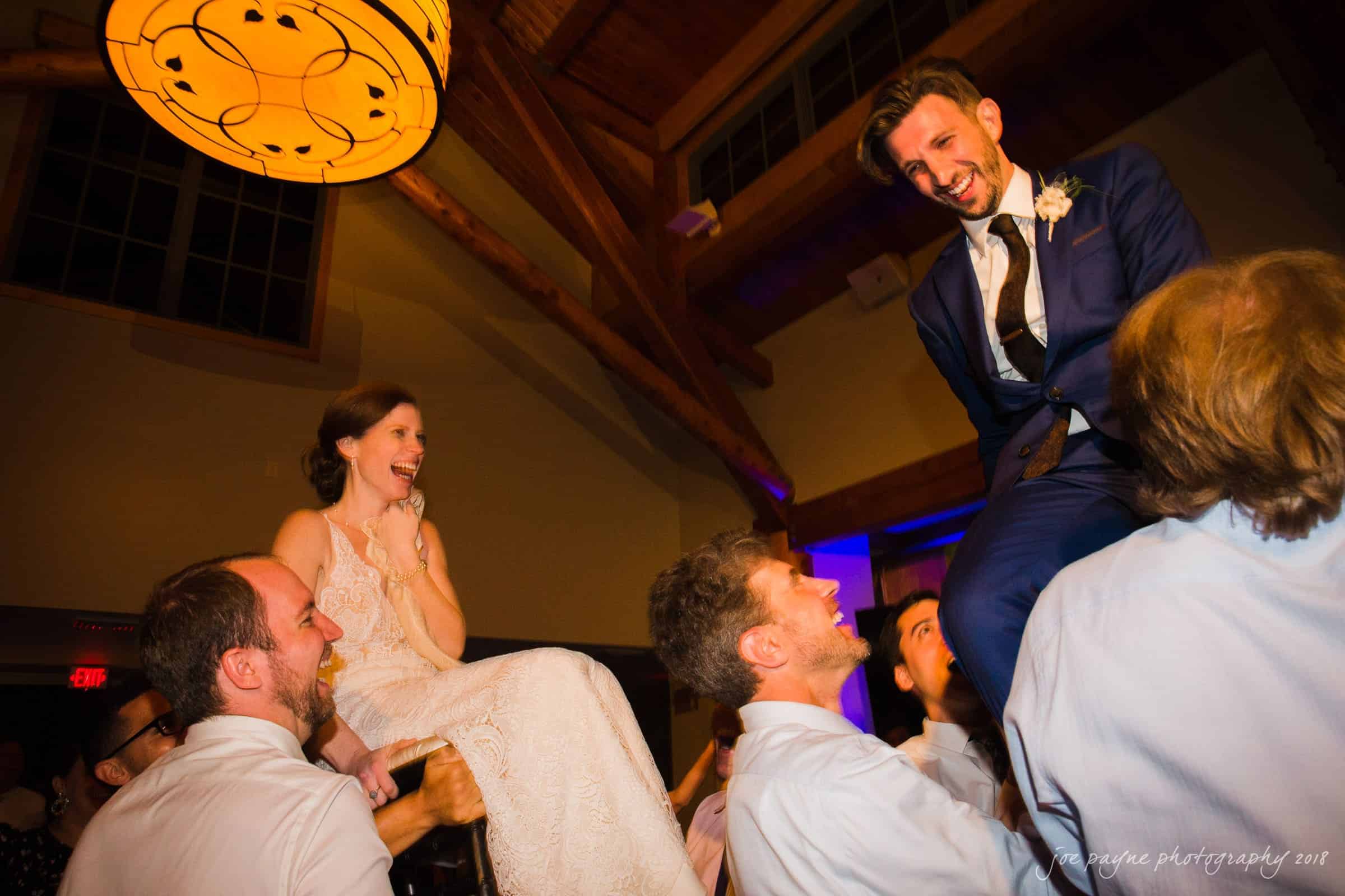 A bride in a white gown and a groom in a blue suit are joyfully lifted on chairs by guests at a wedding celebration. The venue has wooden beams and a large circular light fixture. Both are smiling and laughing.