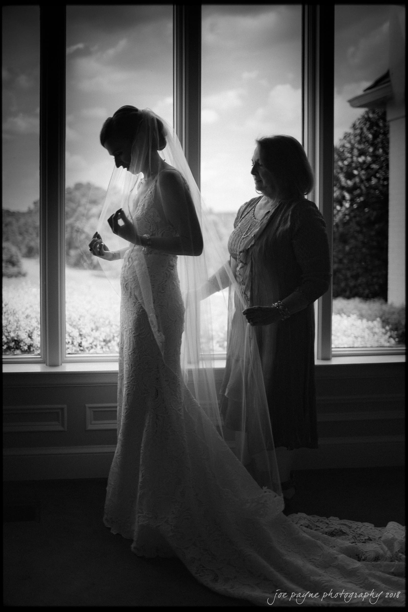 A bride in a lace gown stands by a large window, her veil gently adjusted by another woman beside her. The scene is backlit, casting soft shadows, with a view of a garden outside. The mood is serene and anticipatory.