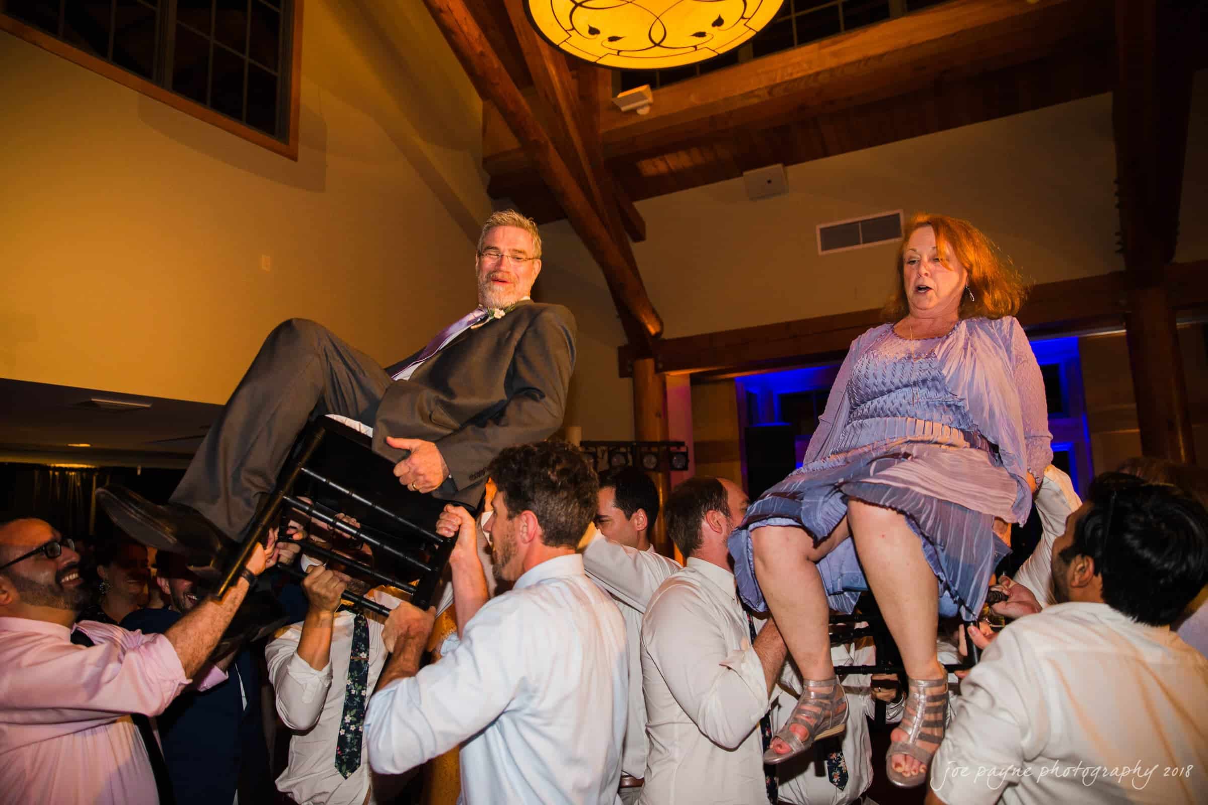 Two people are lifted on chairs by a group of men in a lively dance at a celebration. The room has wooden beams and warm lighting. The people on the chairs are smiling and appear to be enjoying the festive atmosphere.